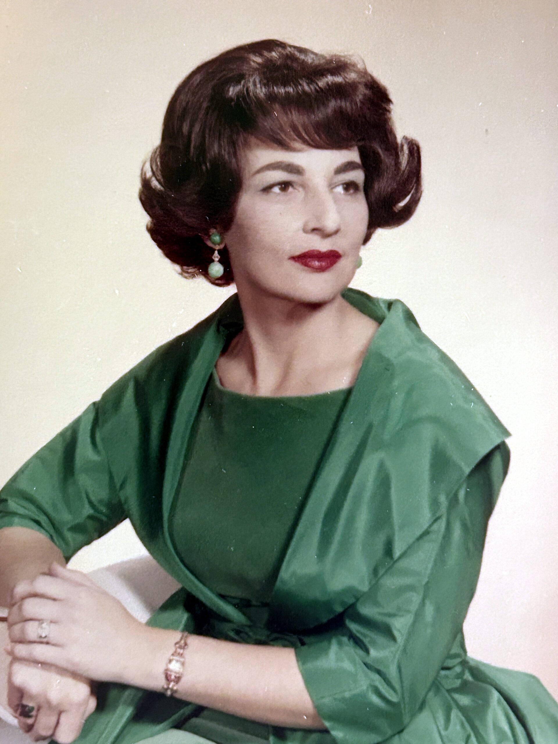Portrait of a woman with short dark hair in a green dress, seated with hands folded and looking to the side.