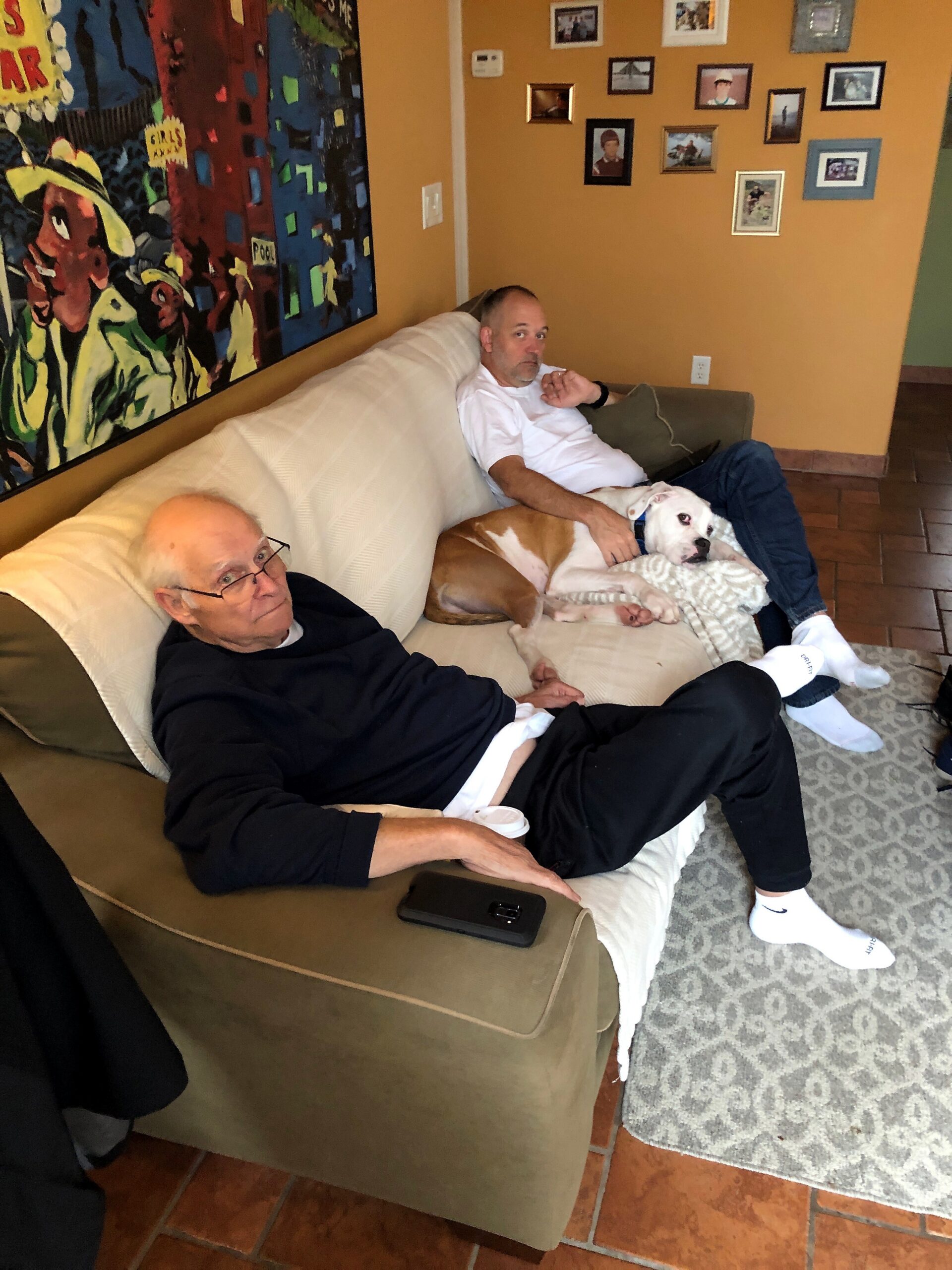 Two men relaxing on a beige sofa in a colorful living room, with a dog snuggled on one man's lap and a collage of photos on the wall behind them.
