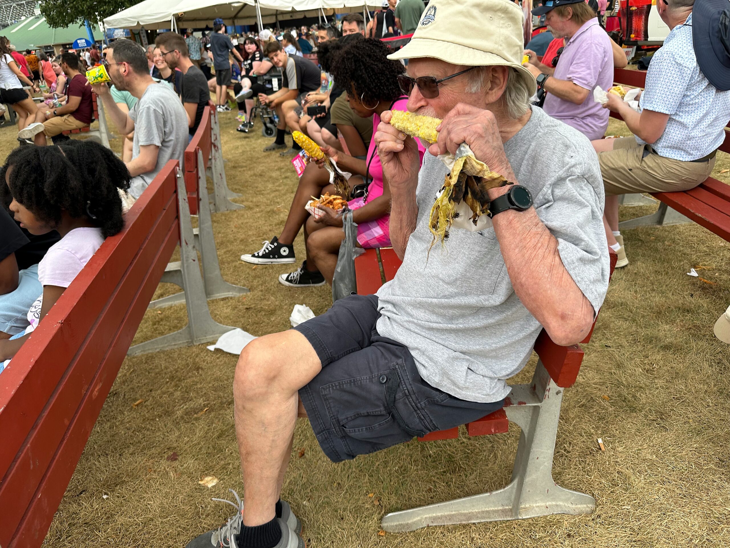Older man in a beige bucket hat and sunglasses eats corn on the cob while seated on red bleachers at a crowded outdoor festival.