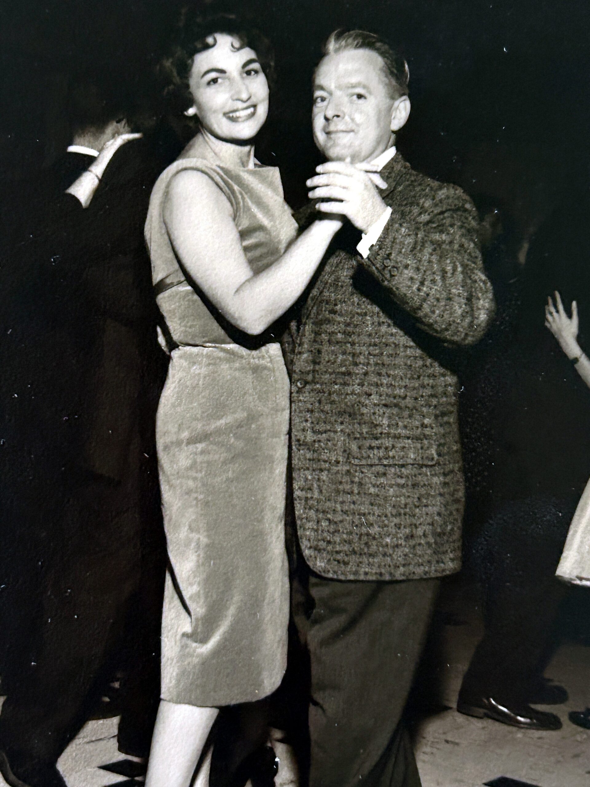 Smiling couple dancing closely at a formal event; woman in a sleeveless dress and man in a textured suit.