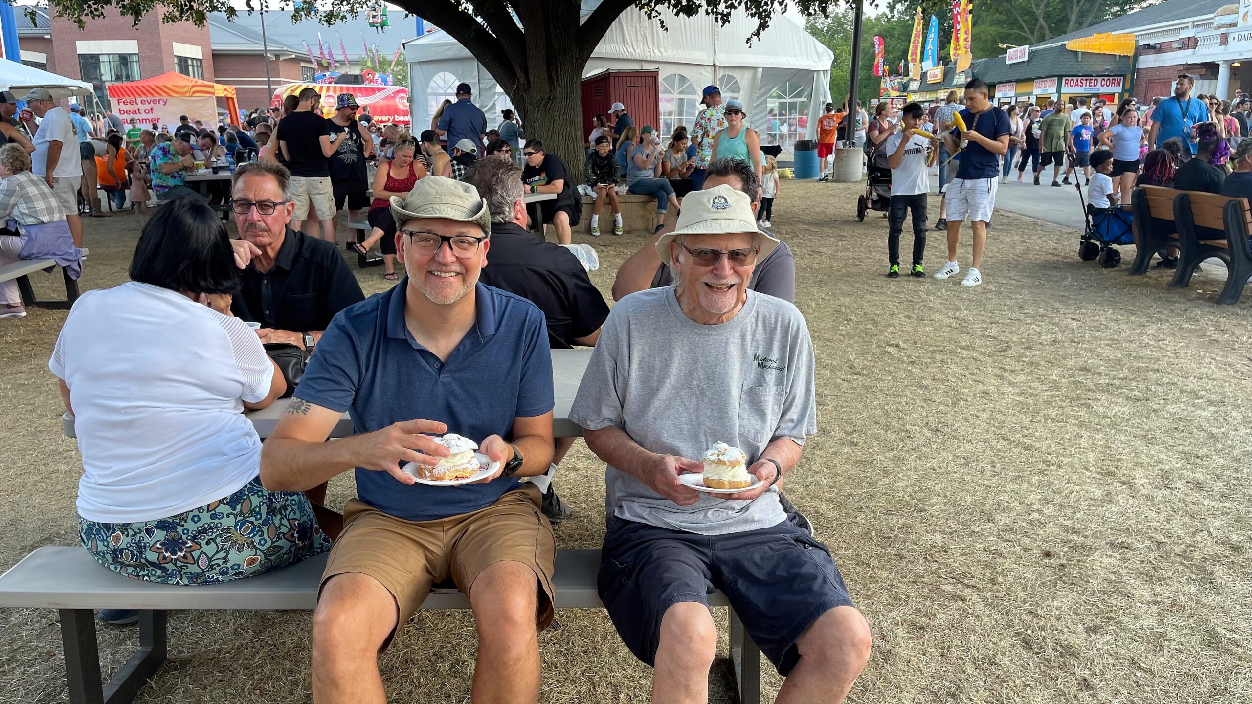 Two men sit at a picnic table under a tree, smiling and eating frosted pastries at a crowded outdoor festival.