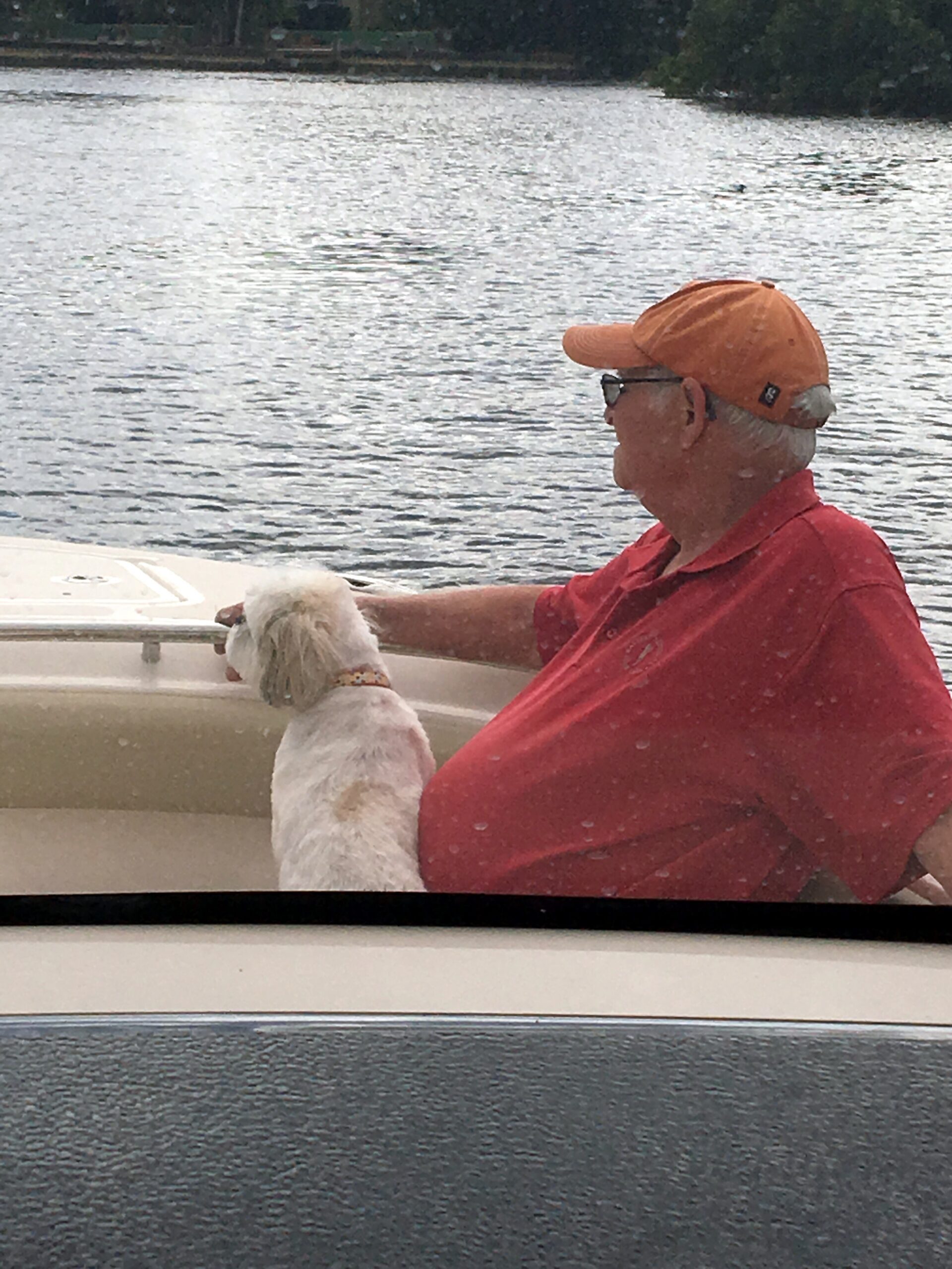 Older man in an orange cap and red shirt sits in a boat with a small white dog by the water.