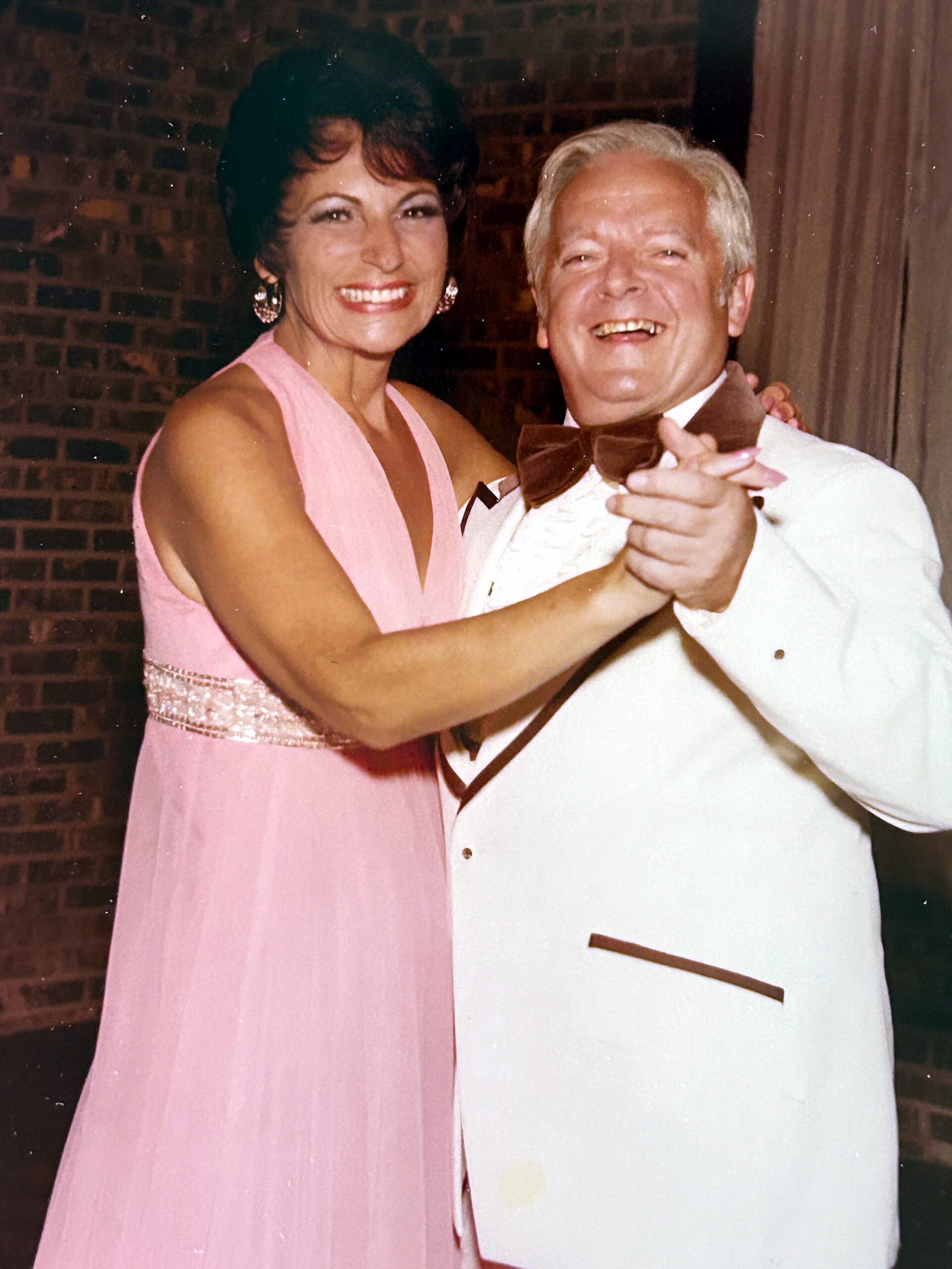 Smiling couple dancing together; woman in a pink evening gown with large earrings and the man in a white tux with a brown bow tie.]