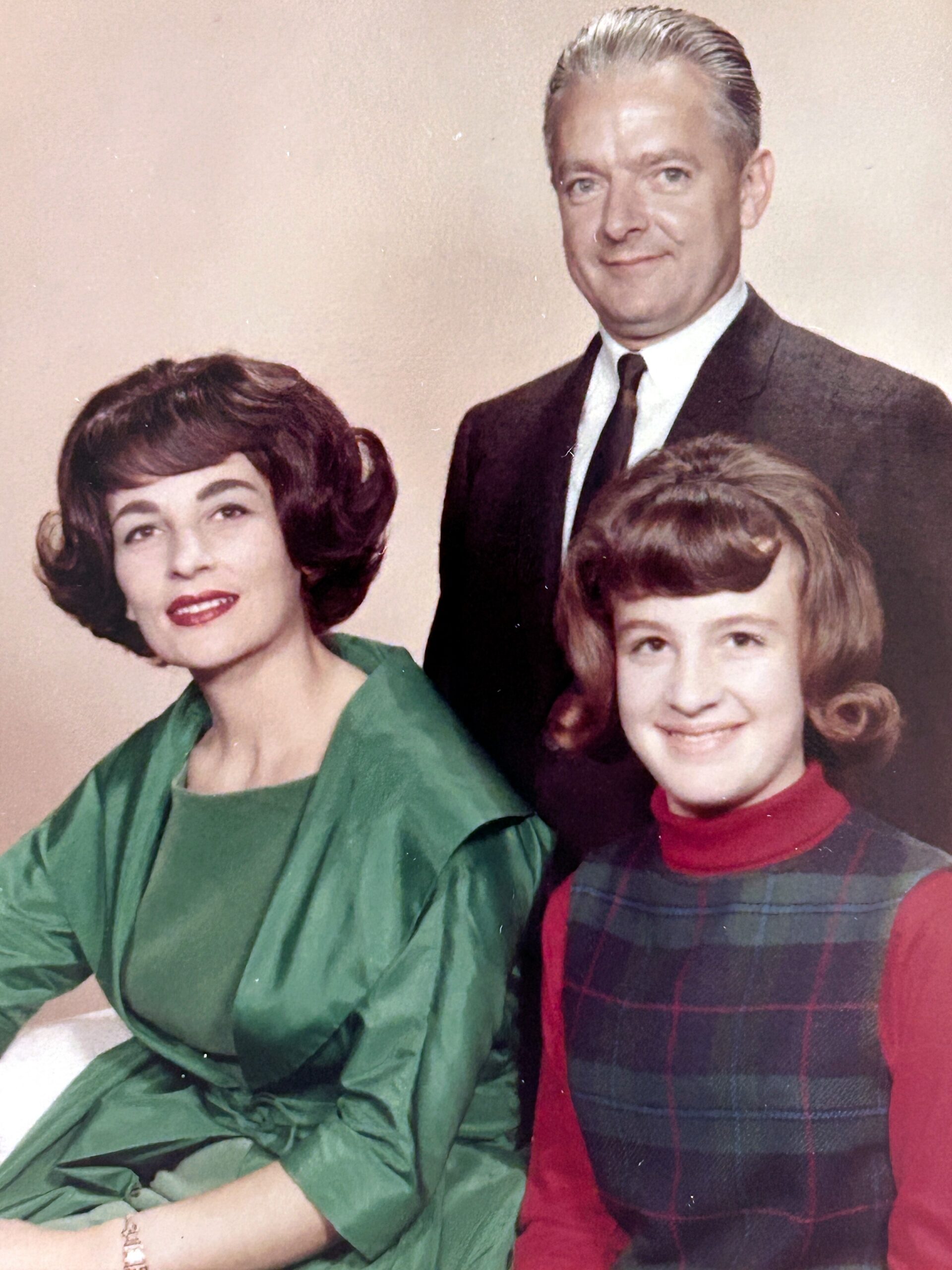 Three family members pose for a vintage portrait: a woman in a green dress, a man in a suit, and a younger girl in a red turtleneck and plaid dress, all smiling.