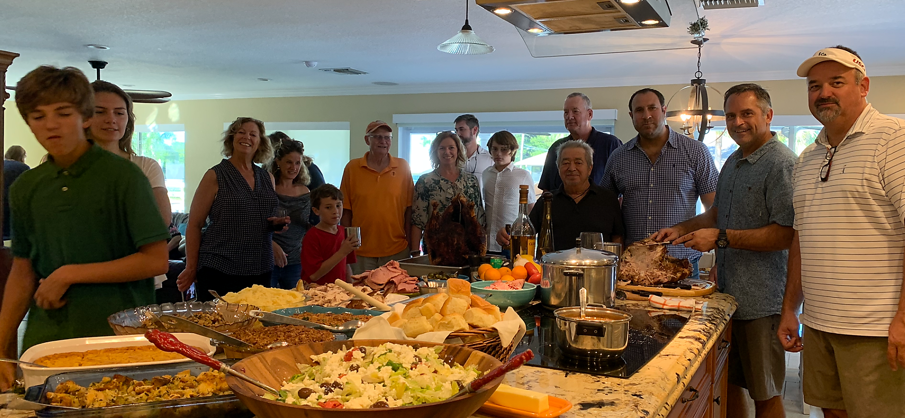 Group of adults and kids gathered around a kitchen island with a big feast, including turkey, salads, bread and bowls, smiling and chatting.