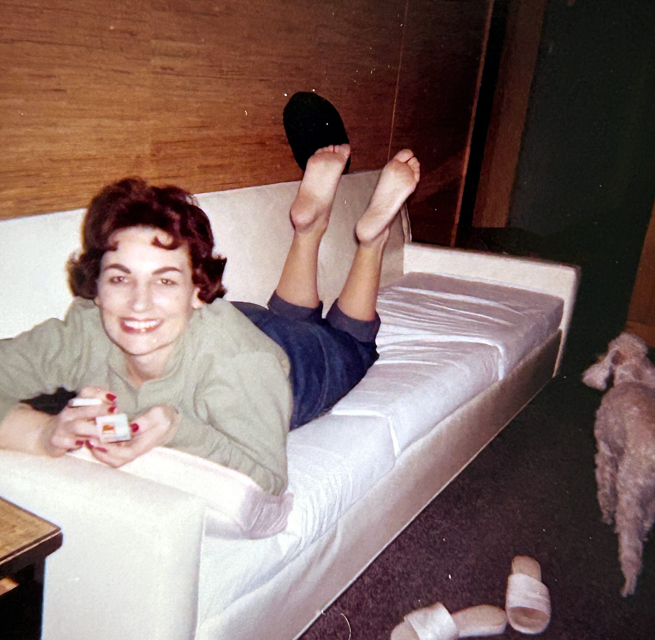 Smiling woman sits on a light sofa with her feet raised, a black slipper resting on one foot and a small object in her hands.