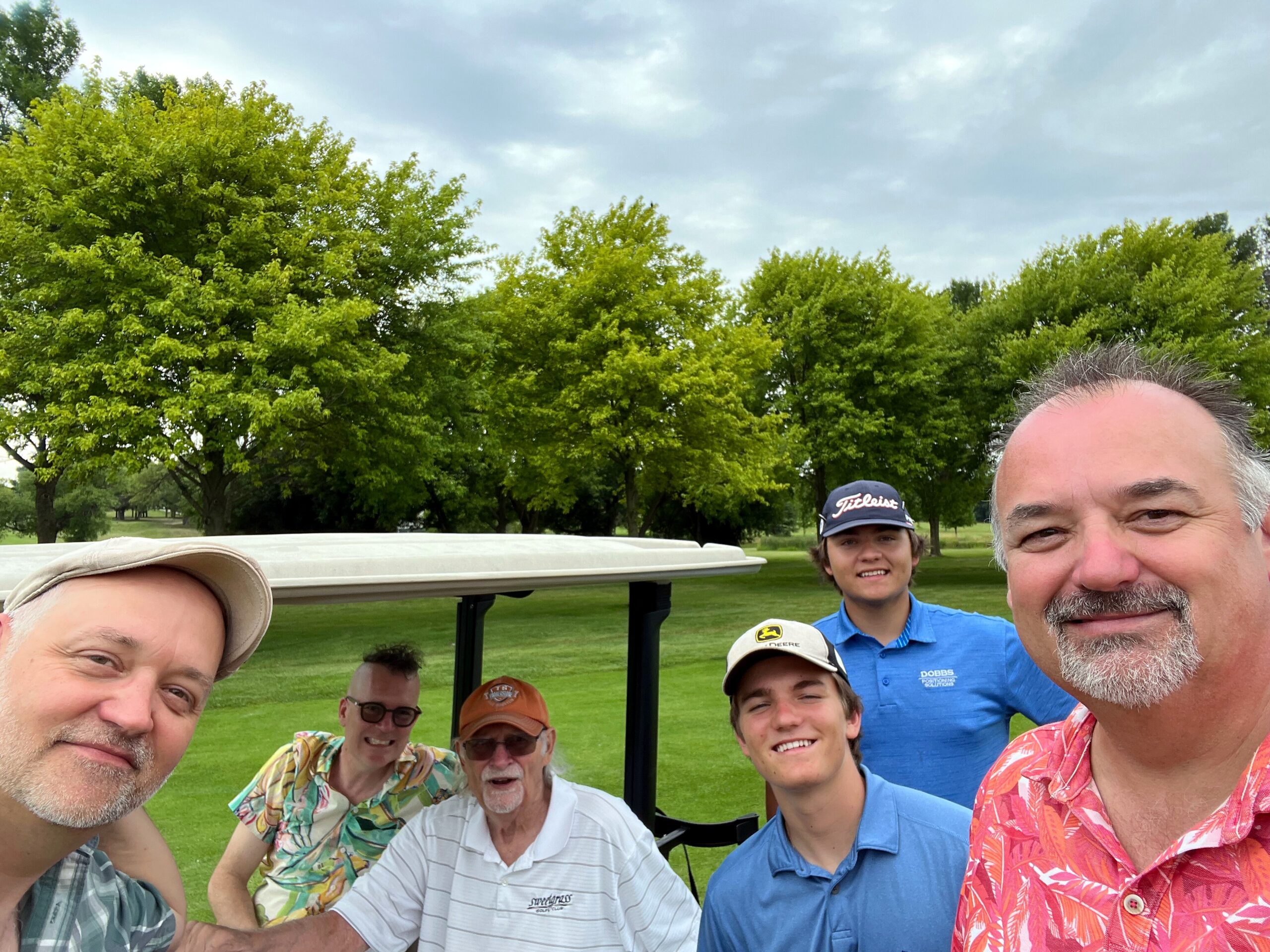 Group of men taking a selfie on a golf course with a golf cart and green trees behind them.