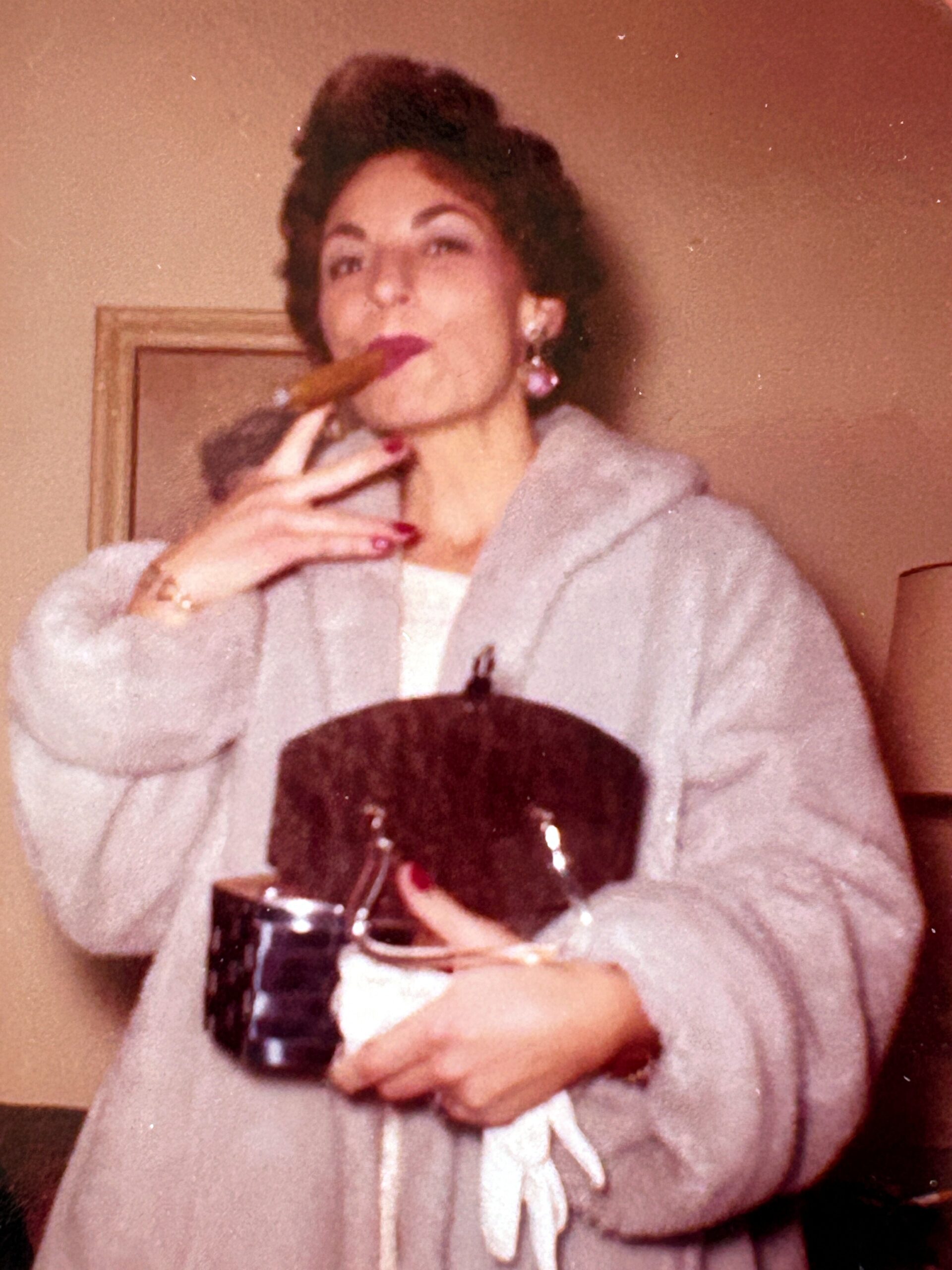 Woman in a pale robe smoking a cigar while holding a wine glass in a cozy indoor setting.