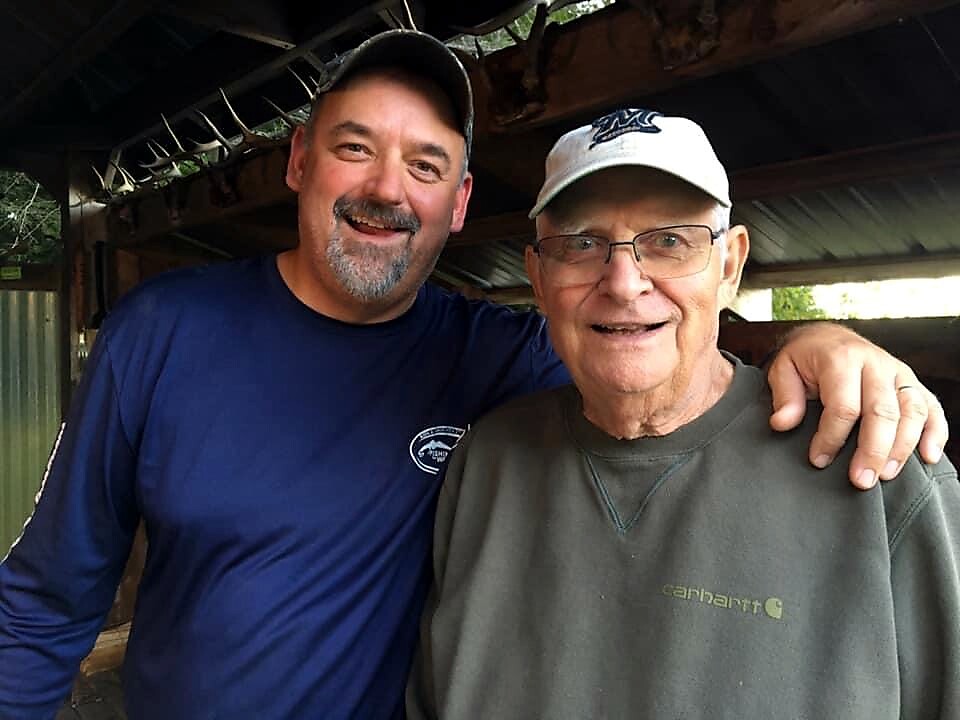Two men smile and pose with arms around each other outdoors under a wooden shelter, wearing caps and casual shirts.