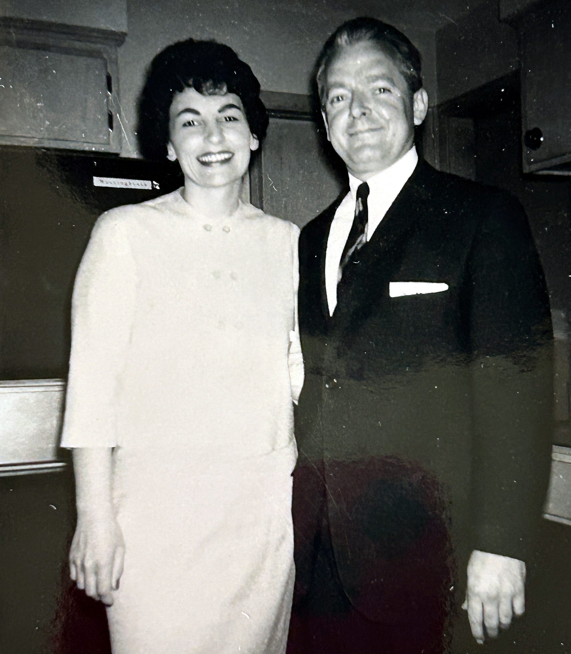 Two smiling adults posing together indoors; woman in light dress on left, man in dark suit on right.