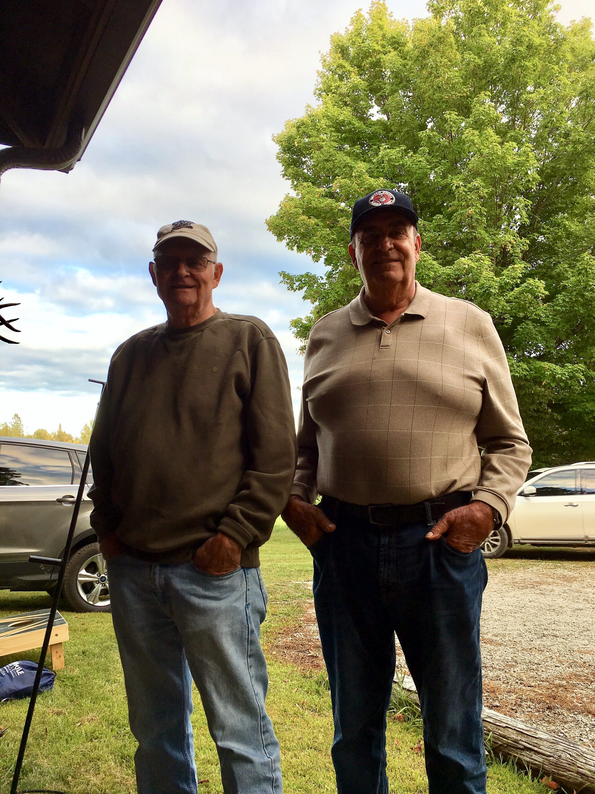 Two older men stand outdoors in casual clothes, smiling, with trees and cars in the background.