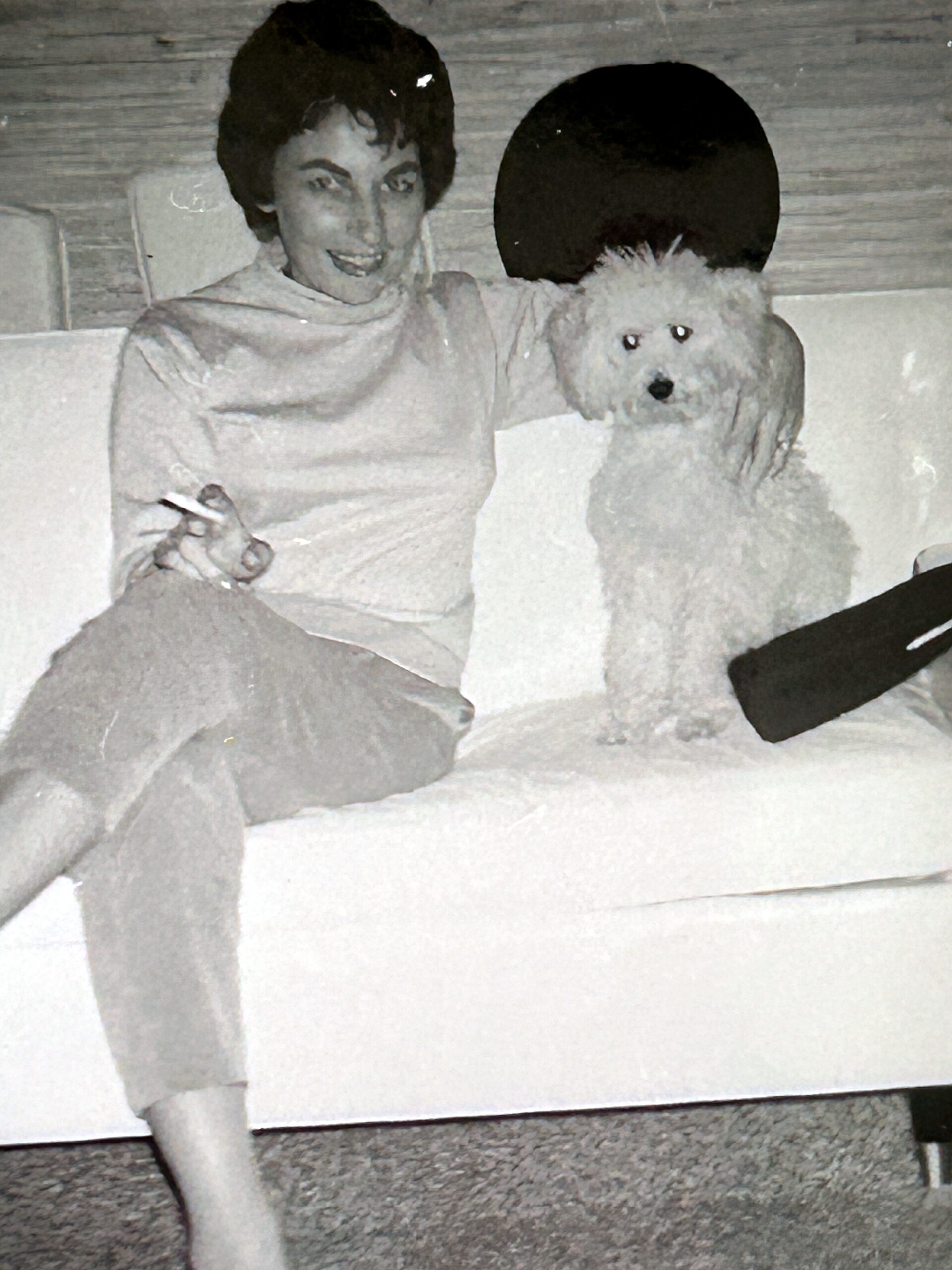 Smiling woman with short dark hair sits on a couch with a fluffy white dog beside her outdoors? indoors.