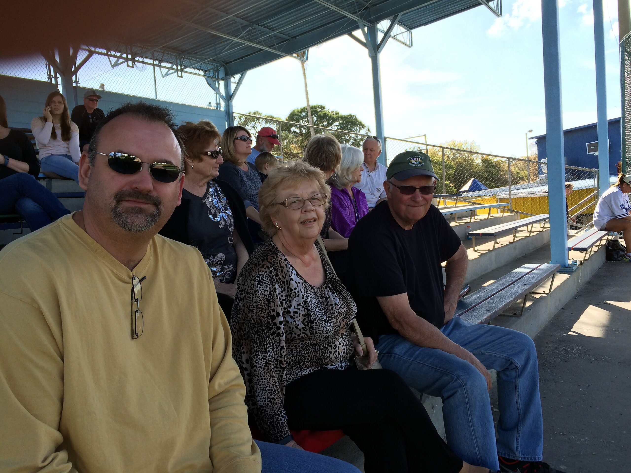 Group of adults seated on blue metal bleachers at an outdoor sports venue, watching an event on sunny day.