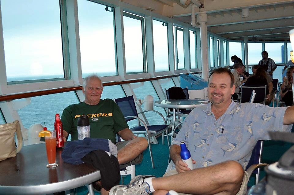 Two men relax at outdoor tables on a cruise ship deck with the ocean visible through large windows, drinks on the table and other passengers in the background.