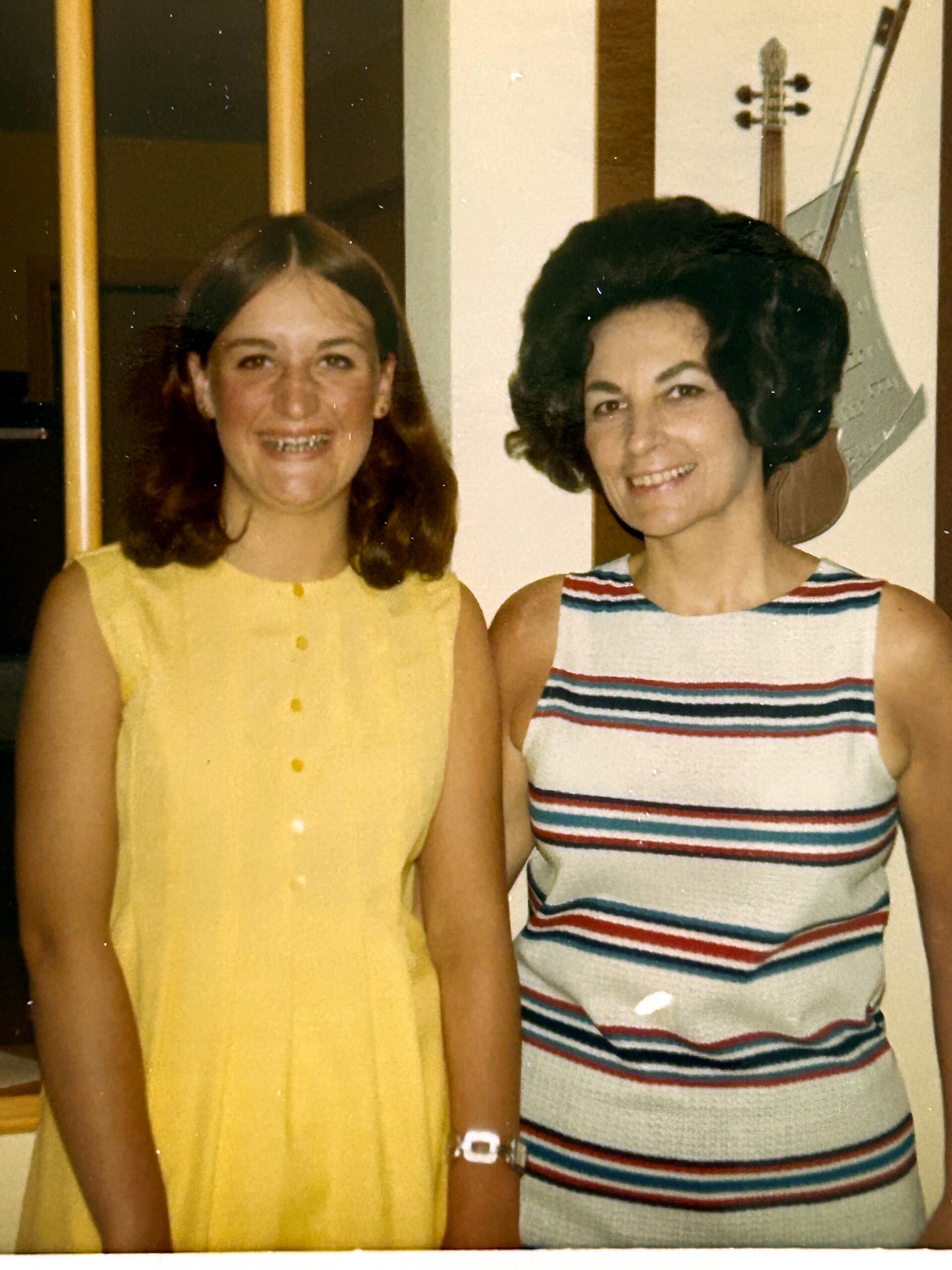 Two women standing indoors, smiling at the camera: left wearing a yellow sleeveless dress with braces, right wearing a striped sleeveless dress, with a guitar visible on the wall behind them.