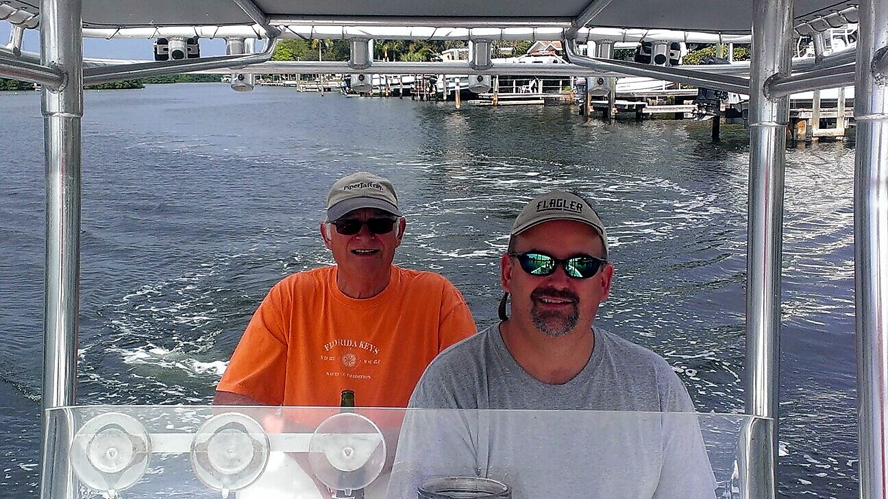 Two men wearing hats and sunglasses sit at the helm of a boat, with a marina and water in the background.