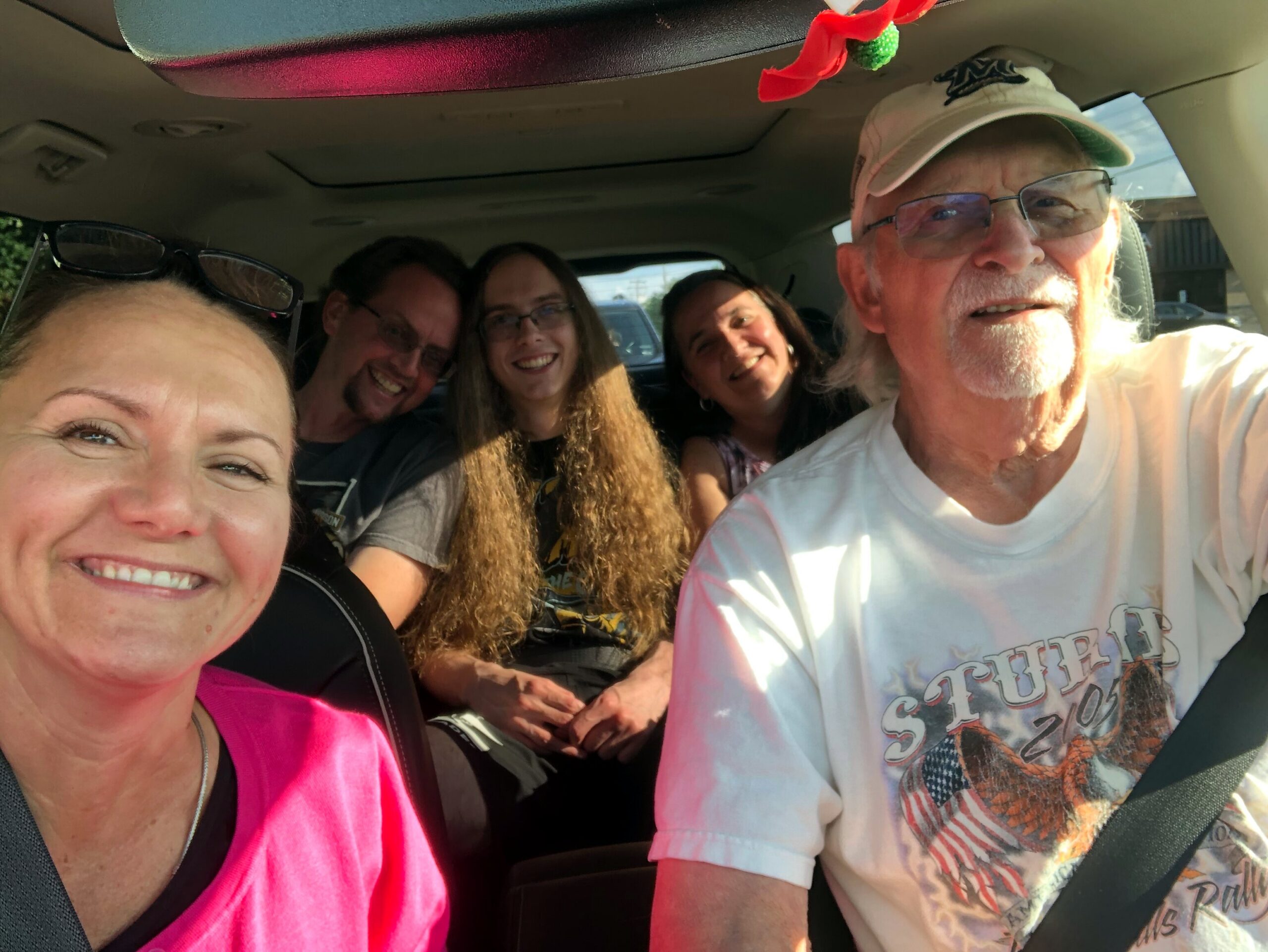 Five adults inside a car smiling for a selfie, wearing seat belts in the front and back seats held close together around the camera.