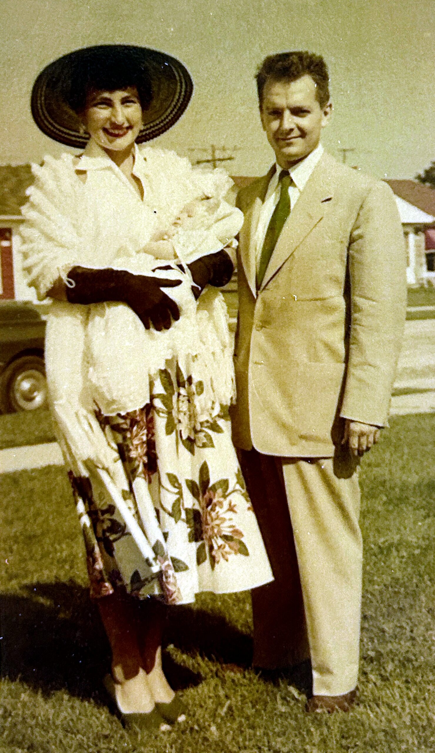 Smiling woman in a white lace dress and large hat holds a baby, standing beside a man in a tan suit outdoors.