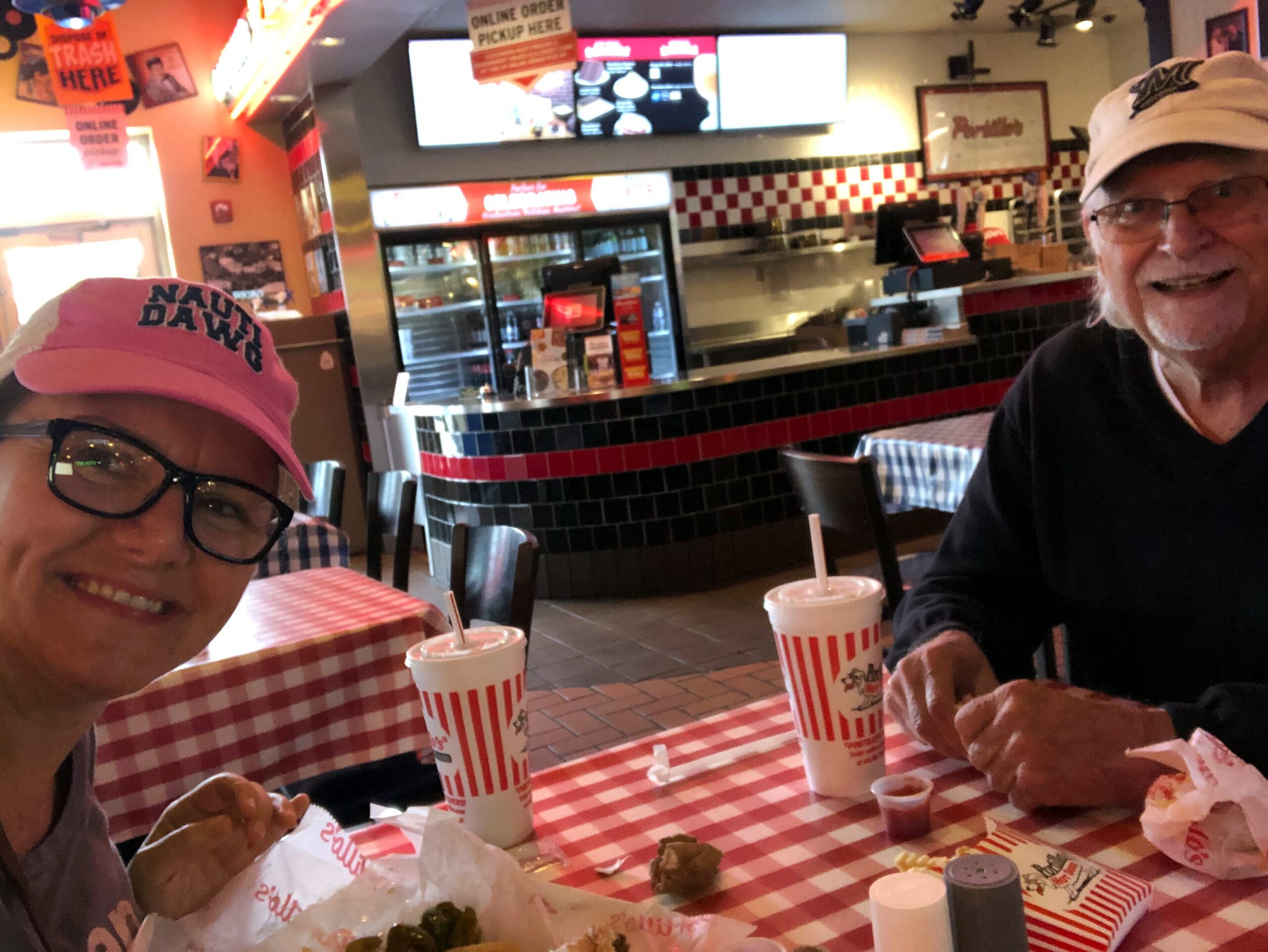 Two smiling people sit at a red-and-white checkered table in a casual diner, with burgers and striped cups in front and a fast-food counter in the background.