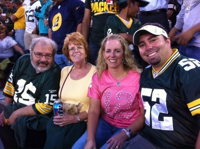 Four Green Bay Packers fans posing in the stadium: a man wearing jersey 15, two women in yellow and pink shirts, and a man in jersey 52 on the right.