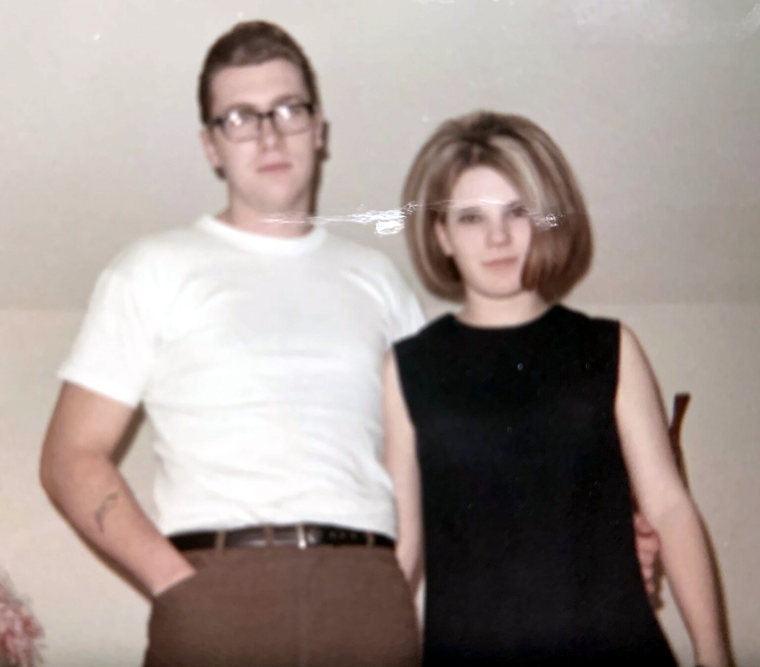 Man wearing a white T-shirt and brown pants stands with a woman in a black dress indoors, posing for the photo.