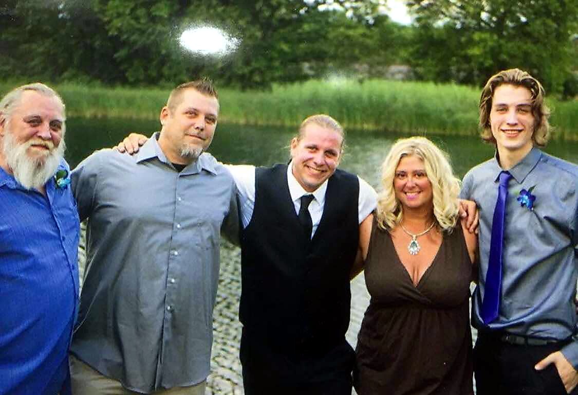 Five adults standing side by side with arms around each other by a lake, smiling outdoors at a sunny day.