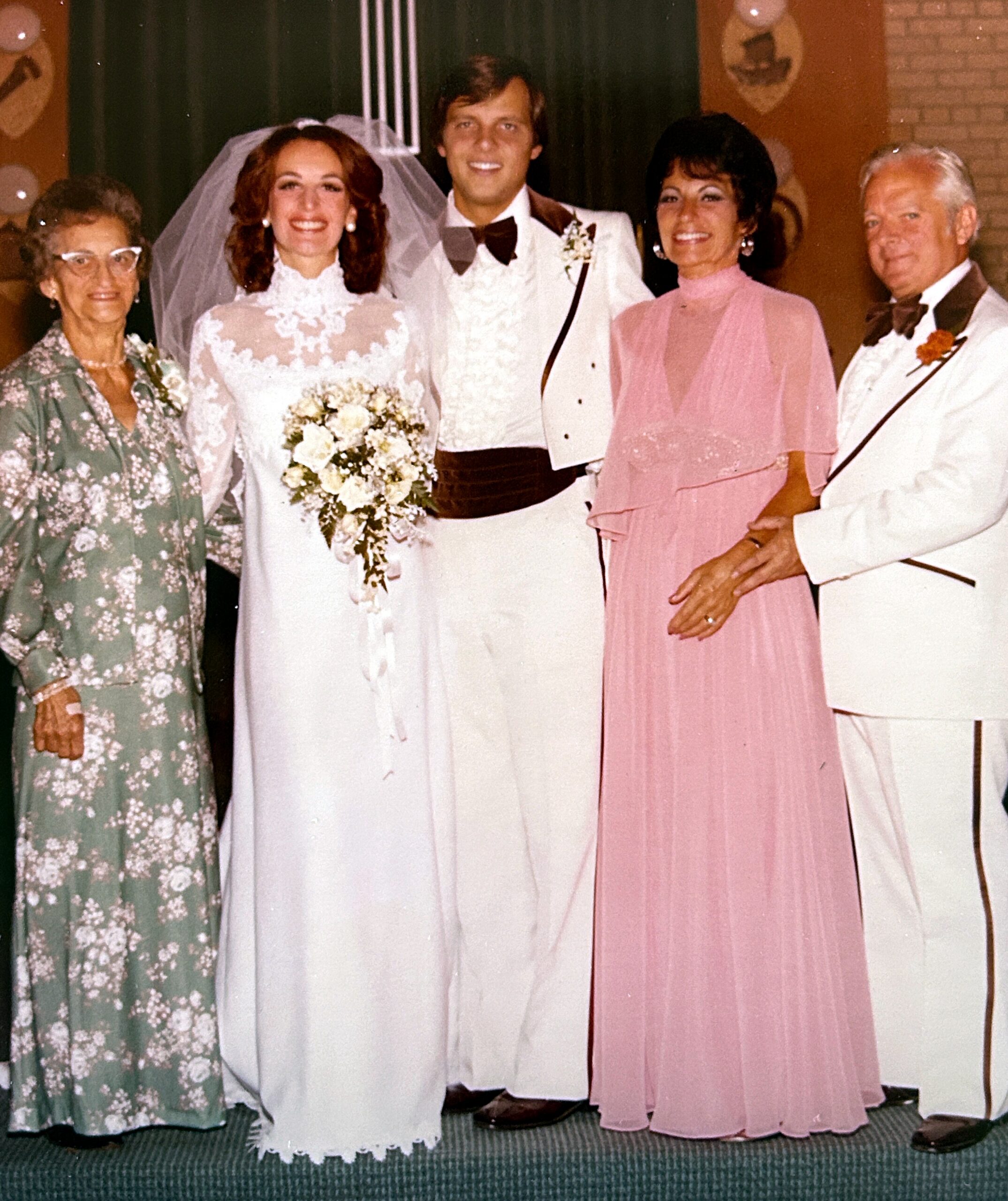 Wedding party photo: bride in lace gown with bouquet and groom in white suit beside two well-dressed guests in pastel outfits.