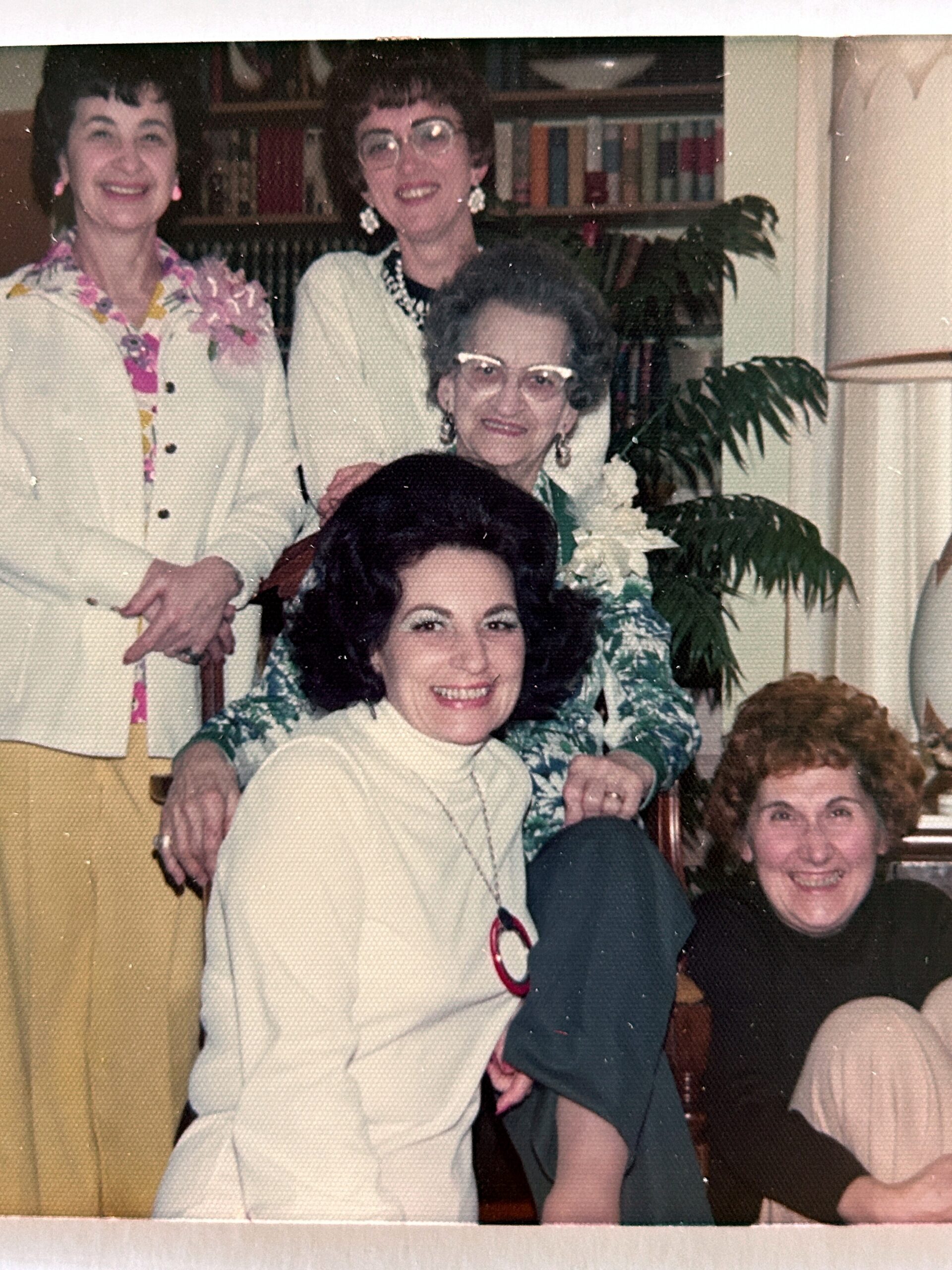 Four women posing together indoors, smiling for a casual group photo; one in a white suit, another in a pale green floral top, with jewelry and short haircuts.
