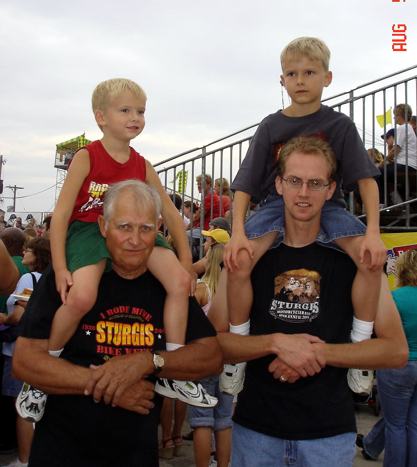 Two men each carry a child on their shoulders at a crowded outdoor festival or event.