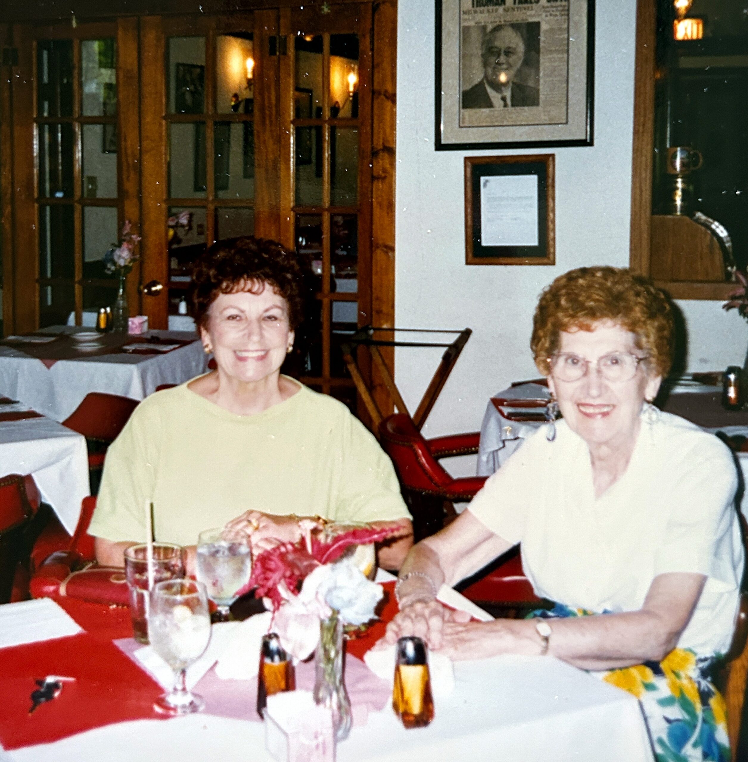 Two smiling elderly women sit at a table during a birthday celebration, with cake and candles in front of them in a restaurant setting.