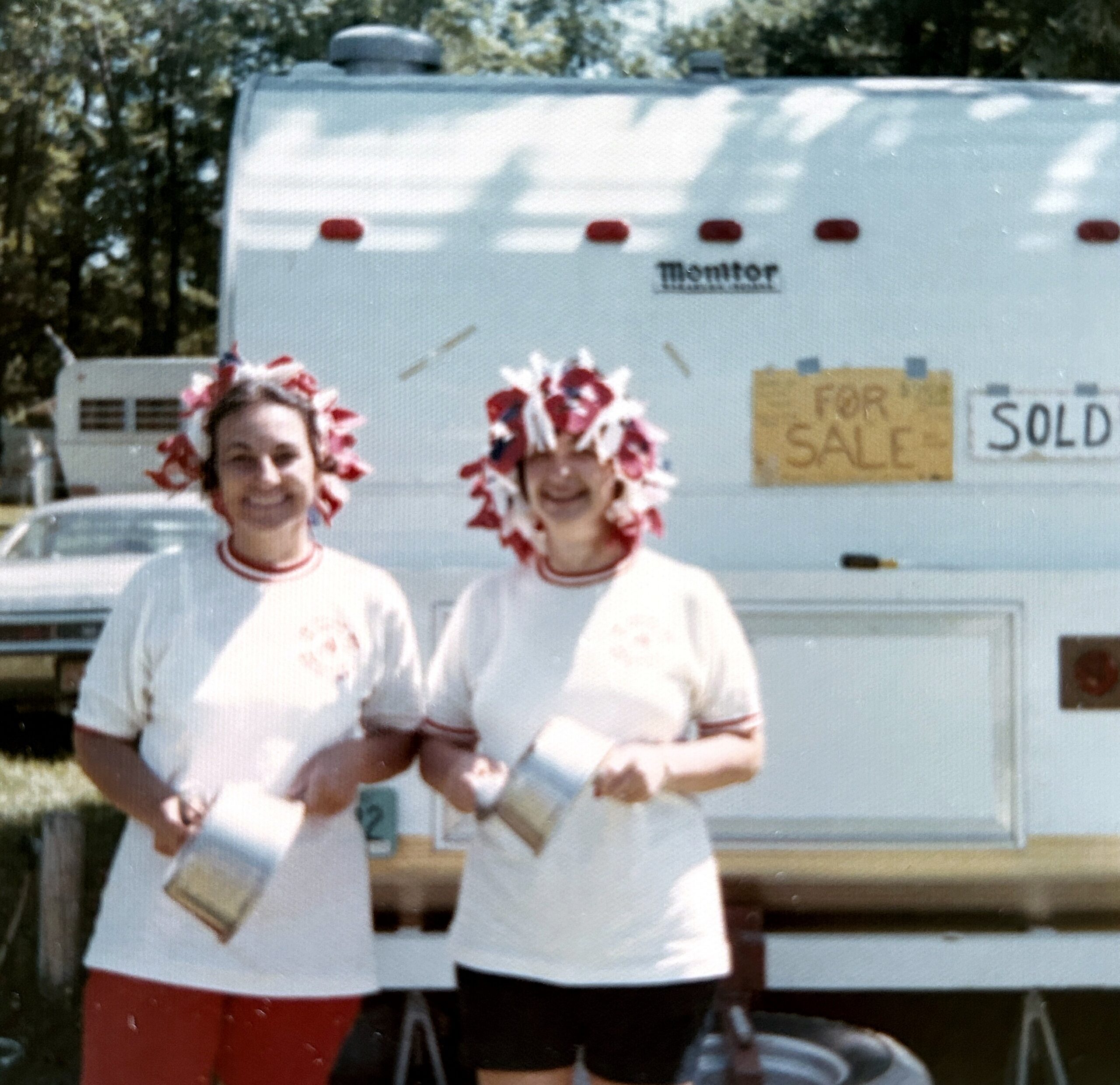 Two cheerleaders in white uniforms with red trim and large red-and-white flower headpieces pose in front of a white trailer with sale signs visible.