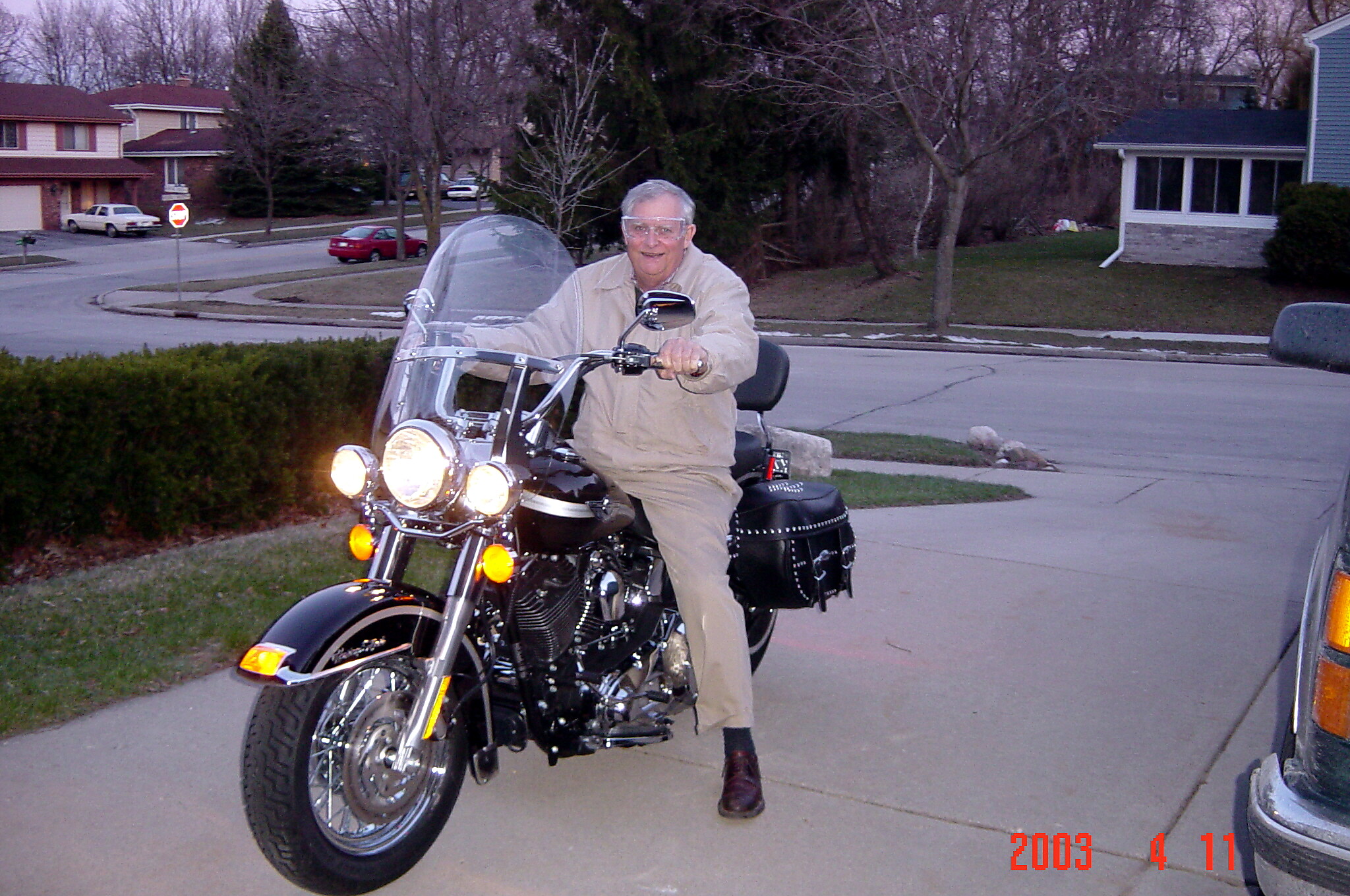Older man in a beige jacket and goggles rides a black motorcycle down a suburban street with a large windshield.