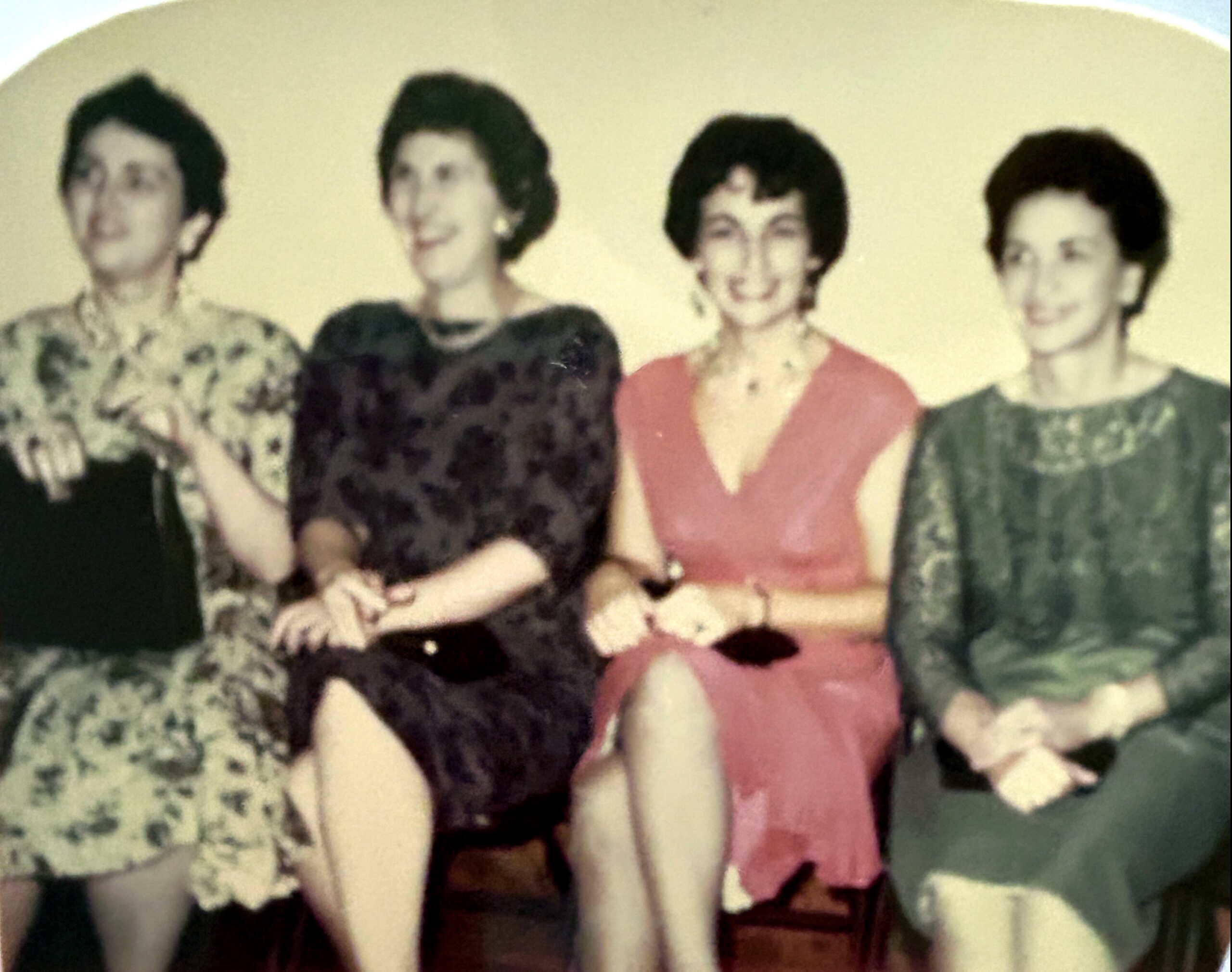 Group of four women standing together, smiling, wearing vintage dresses in a posed photo (1960s style).