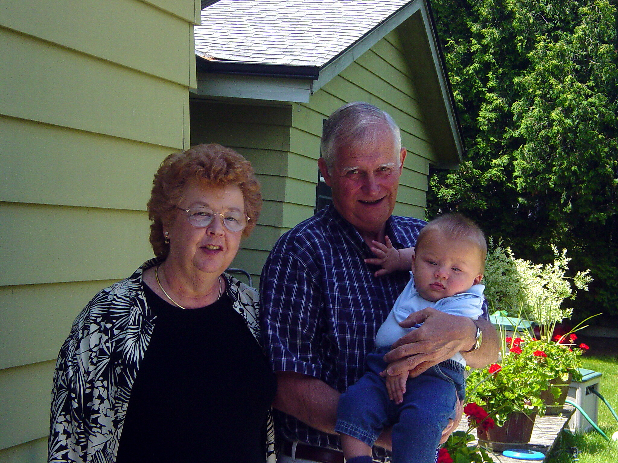 Smiling elderly woman and man holding a baby boy outdoors in front of a green house with plants behind them. (family photo)