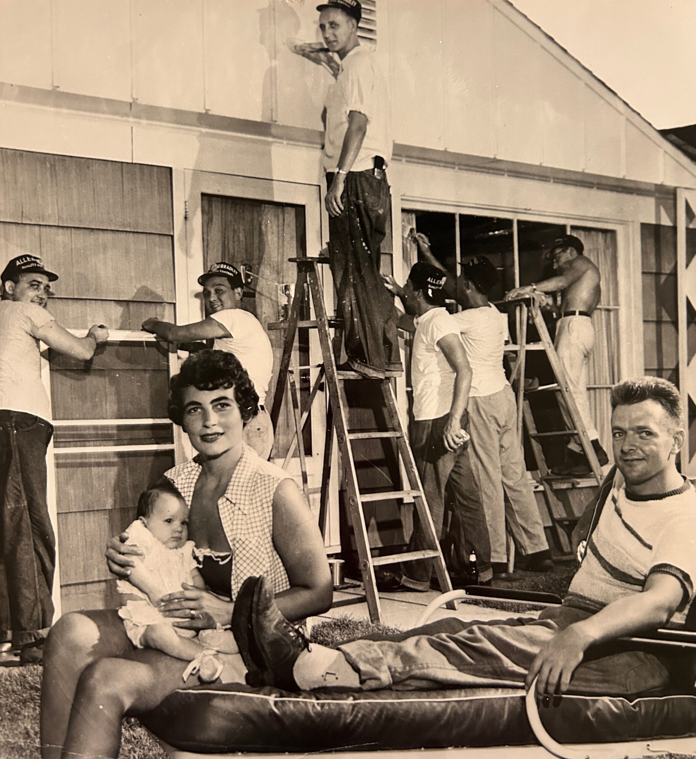 Vintage photo of a construction crew gathered on a ladder and platform against a building; a woman with a baby sits in the foreground while others work.
