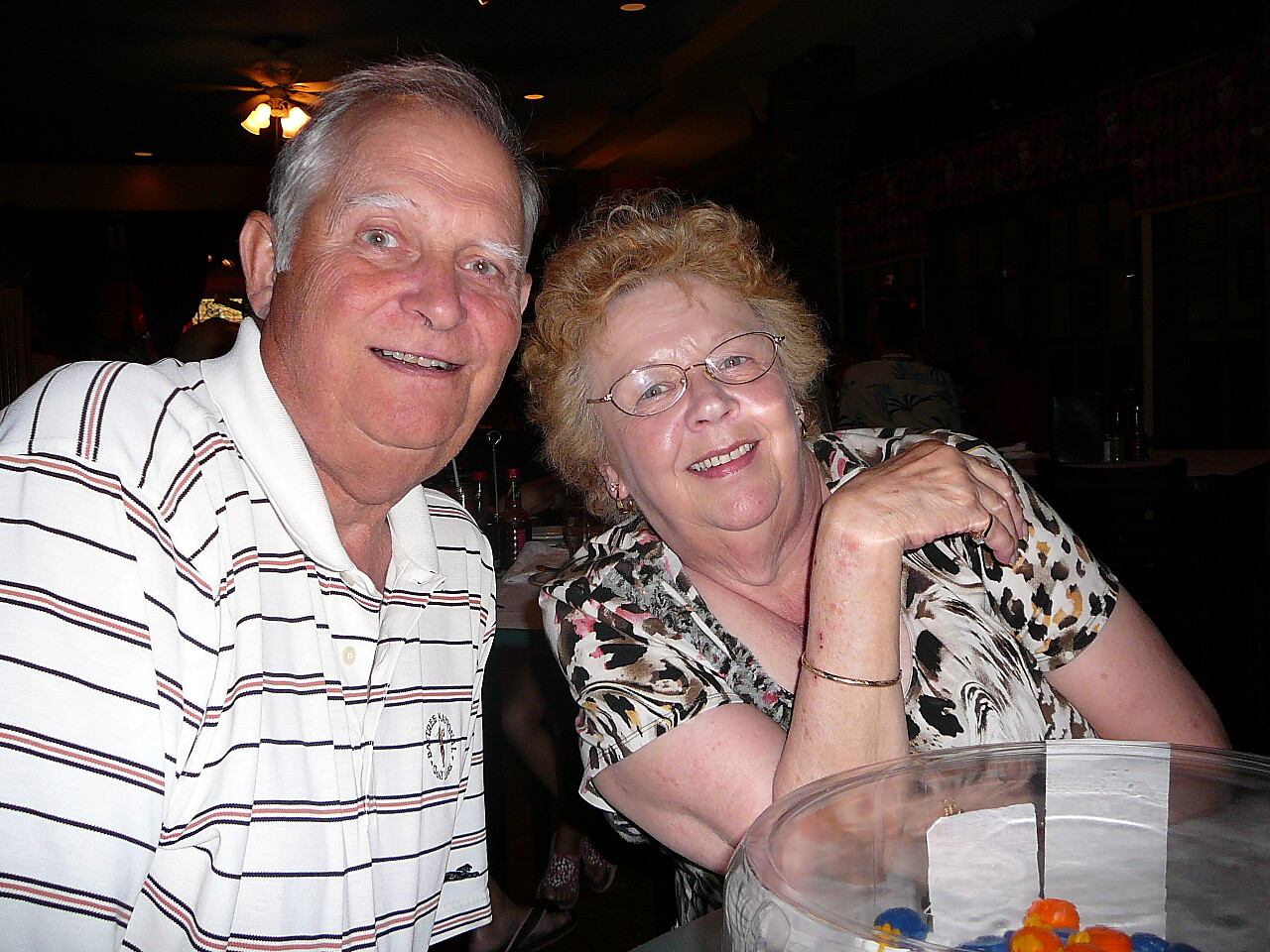 An elderly couple smiling at a restaurant table, the man in a striped polo and the woman wearing glasses leaning close together near a clear bowl with colorful candy.