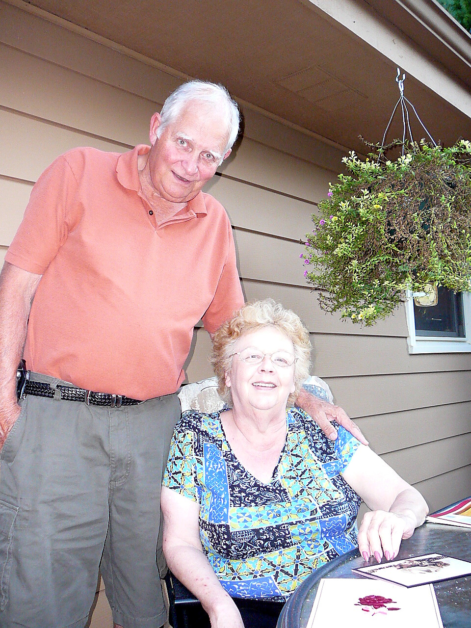 Older man in an orange polo stands behind a smiling seated woman on a porch, his hand resting on her shoulder.