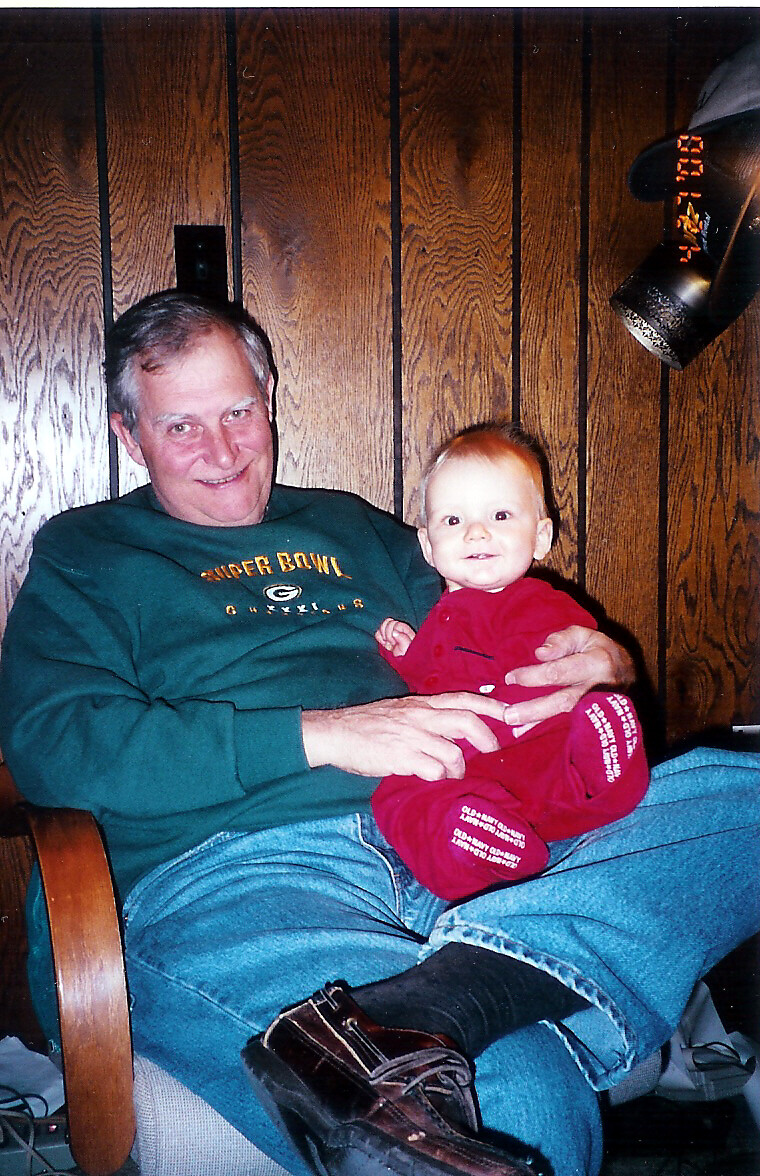 Man in a green sweatshirt sits in a chair, smiling, holding a baby in a red outfit on his lap against wood paneling.
