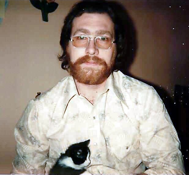 Bearded man with glasses wearing a light patterned shirt, indoors, with a black-and-white cat in his lap.