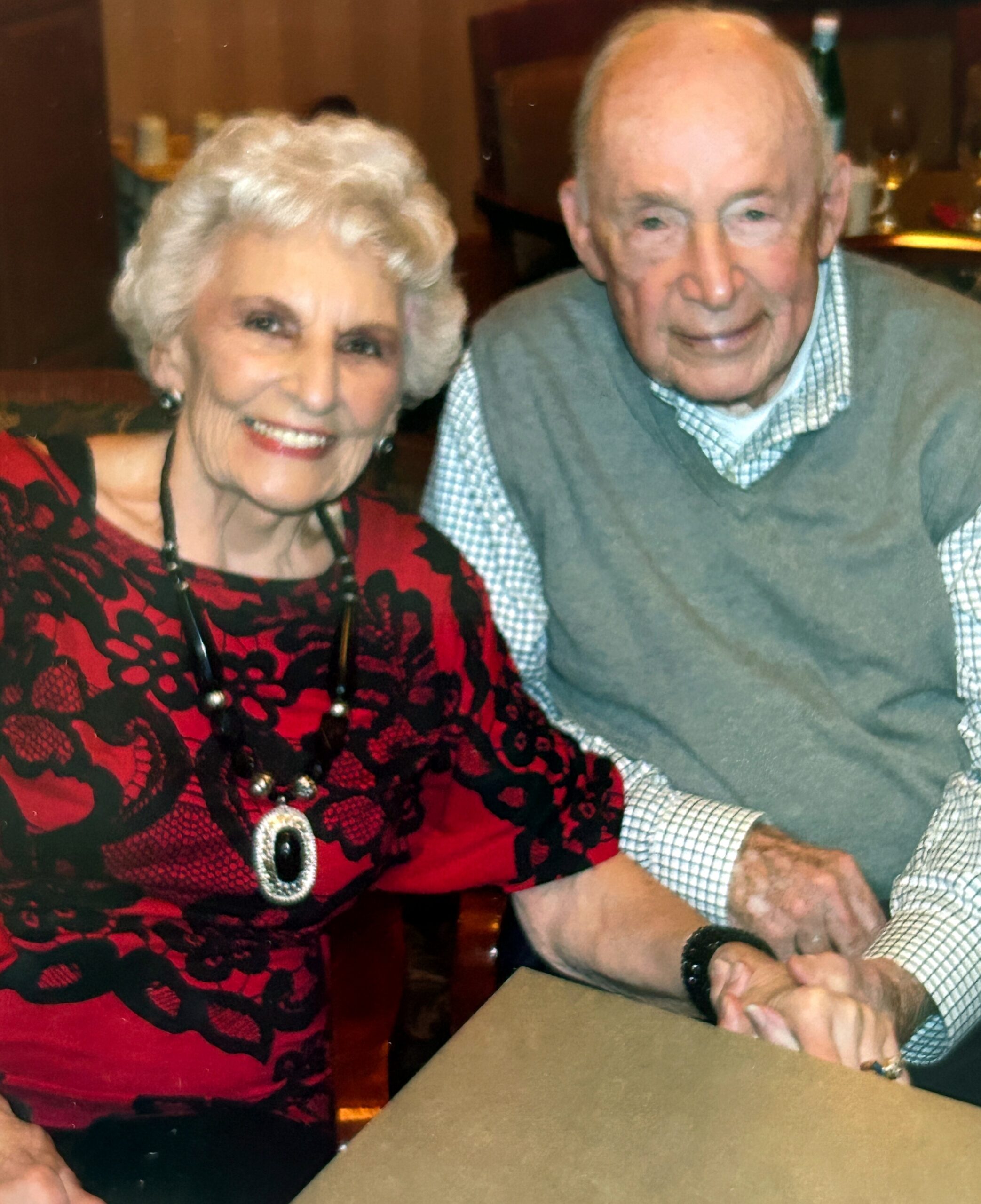Smiling elderly woman in a red and black lace top sits beside an elderly man in a gray sweater at a table, posing for a photo.