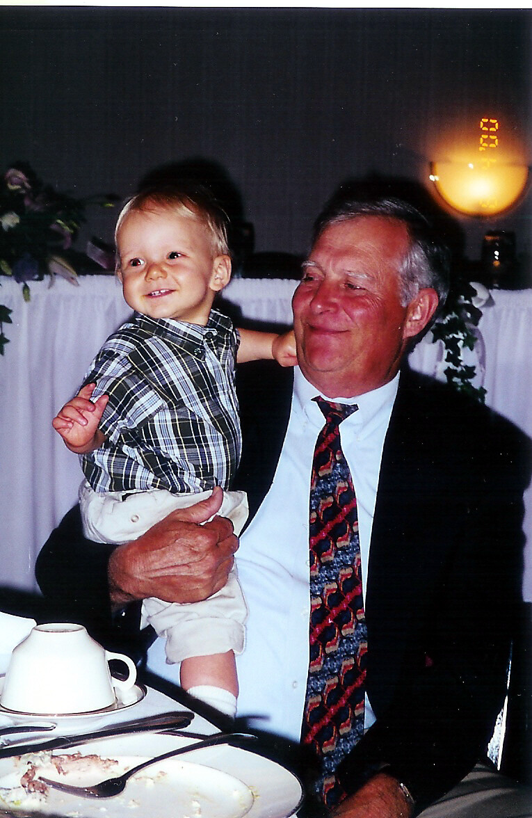 Grandfather in a suit holds a smiling toddler at a banquet-style dinner table with plates and cups visible.