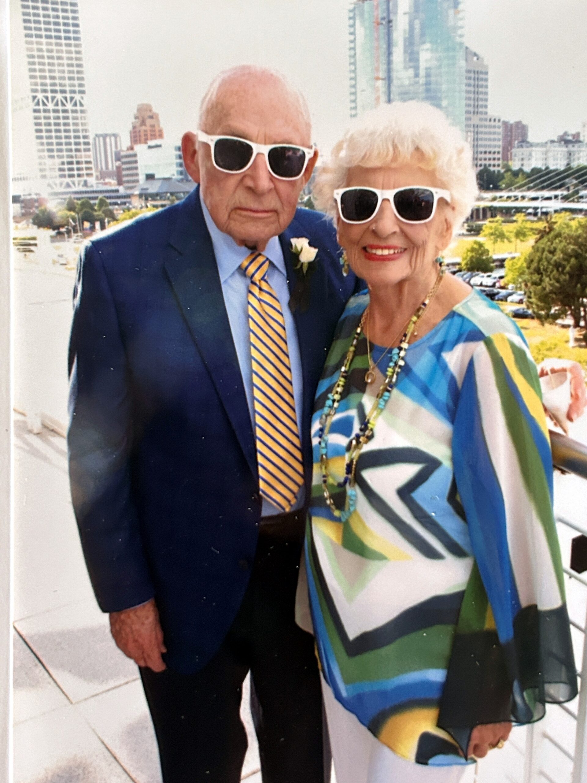 Smiling elderly couple wearing white sunglasses in front of a city skyline; man in a blue suit and striped tie, woman in a colorful blouse.