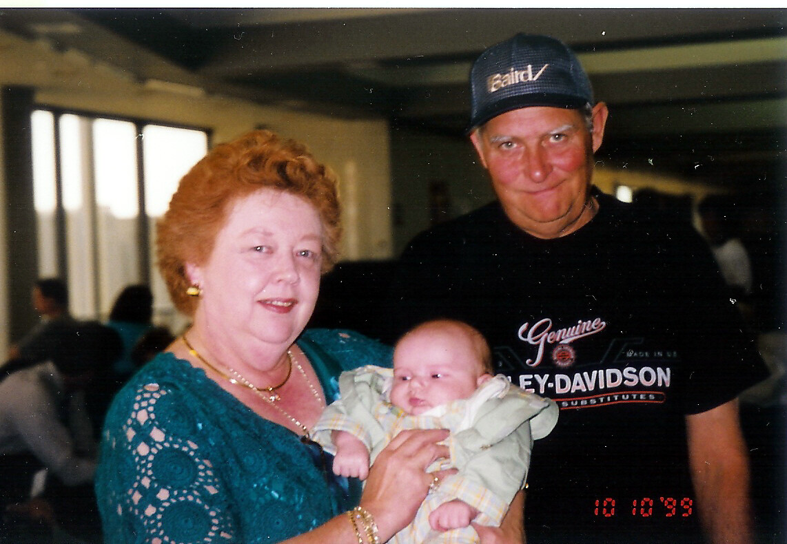Older woman in teal lace holds a newborn baby, with a man in a Harley-Davidson cap beside them indoors.