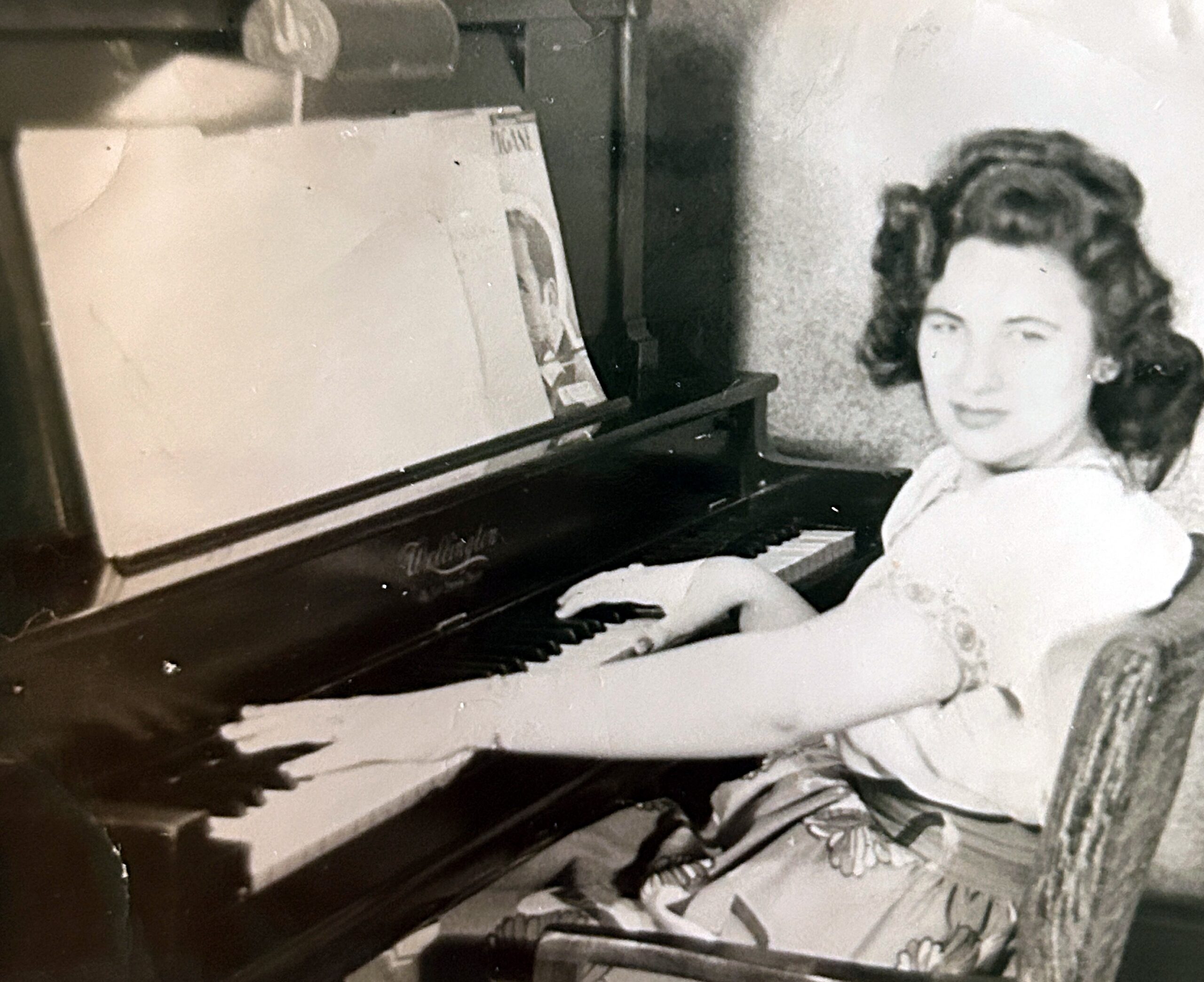 Black-and-white photo of a woman sitting at a grand piano, hands on keys, looking toward the camera.