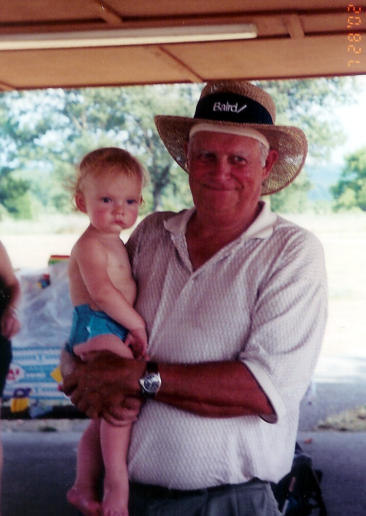Older man in a checkered shirt and straw hat holding a young toddler in blue swim trunks under a shelter at a park.