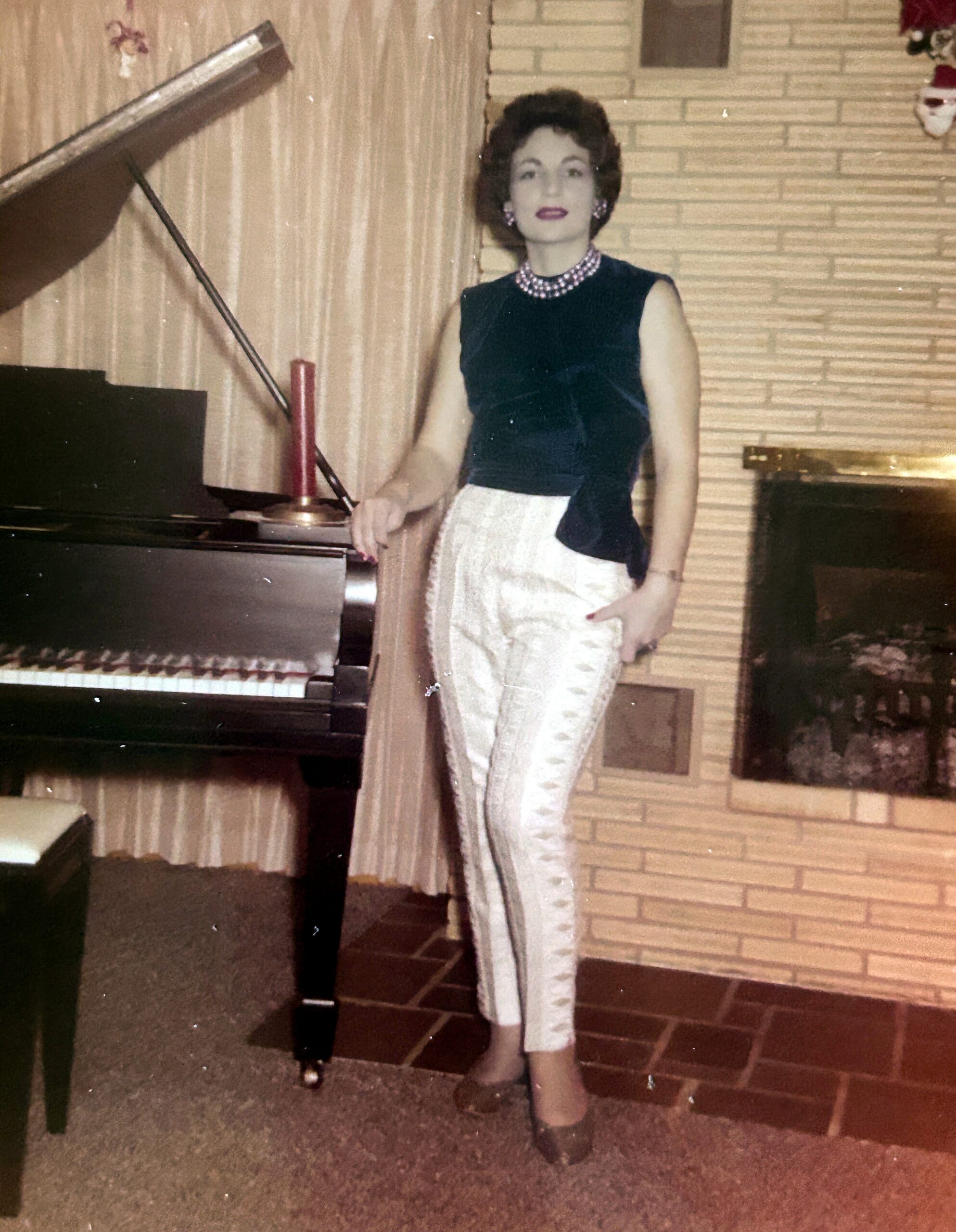Woman in a sleeveless dark velvet top and white skirt with a pearl necklace standing beside a grand piano in a living room.