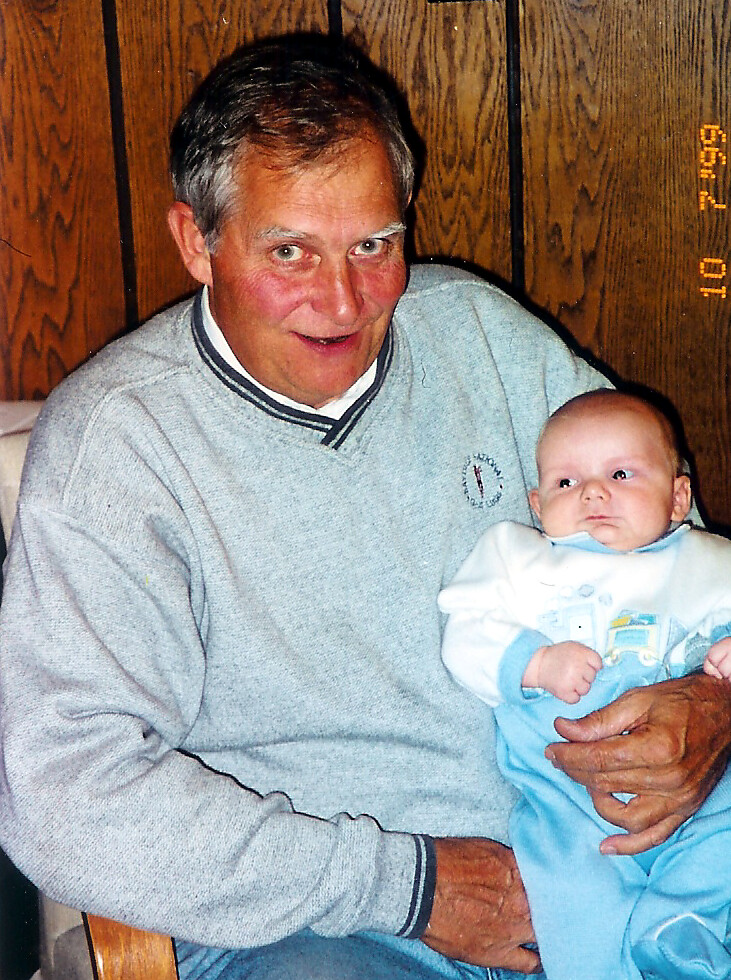 Older man in a gray sweater holding a baby in a blue onesie indoors.](