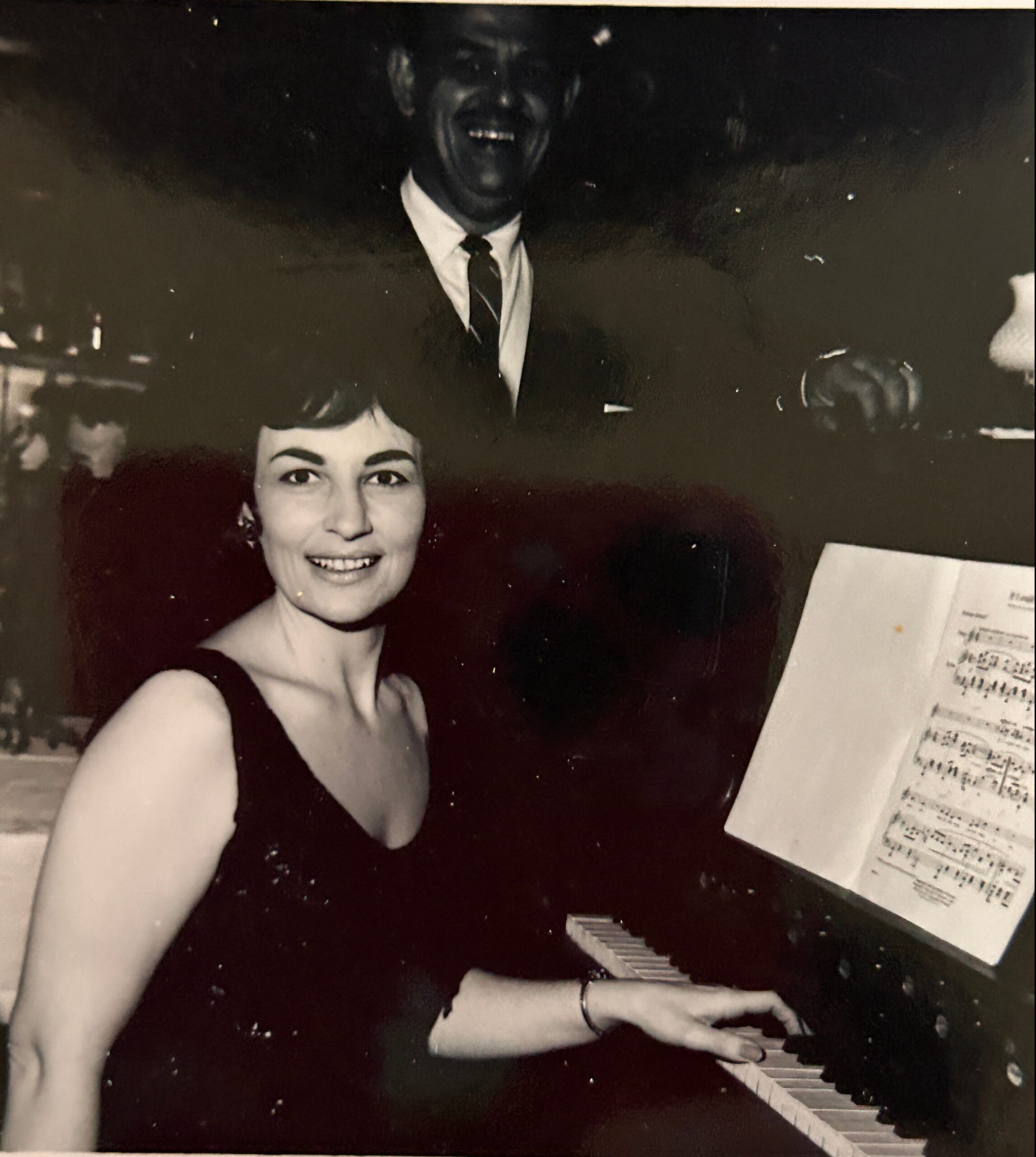 Smiling woman in a black dress with a man in a suit behind her, at a social setting with a piano nearby.