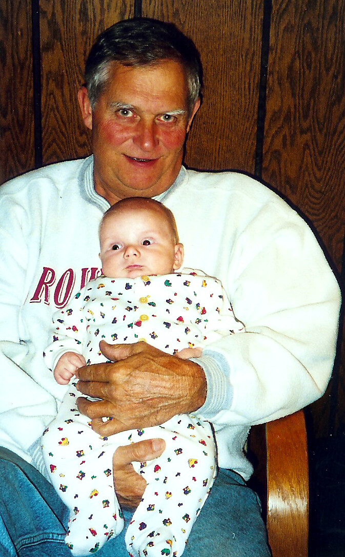 Grandfather in a light sweater cradles a baby in a patterned white onesie, sitting indoors against wood paneling aahana homey scene
