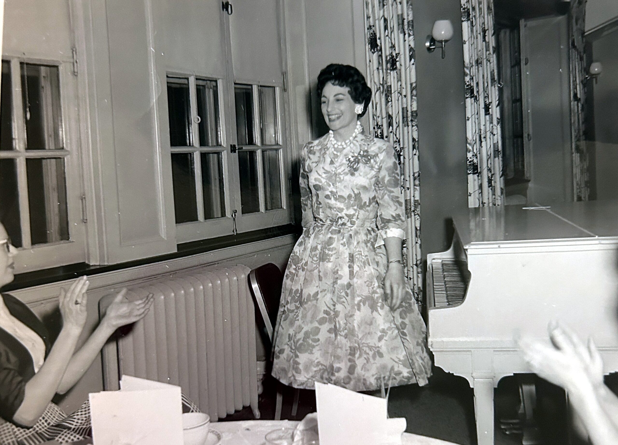 Smiling woman in a floral dress with pearls stands by a piano in a room with patterned curtains; audience claps nearby, vintage photo.